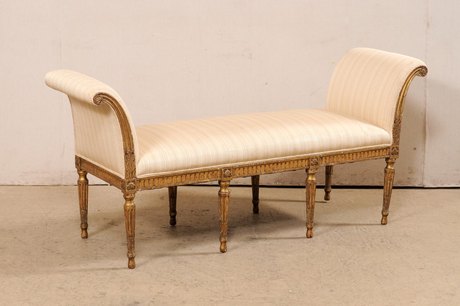 A French Louis XVI style carved wood and upholstered bench from the turn of the 19th and 20th century. This antique bench from France is backless, and is flanked with a pair of gracefully curved arms that terminate into scrolled volutes with floral