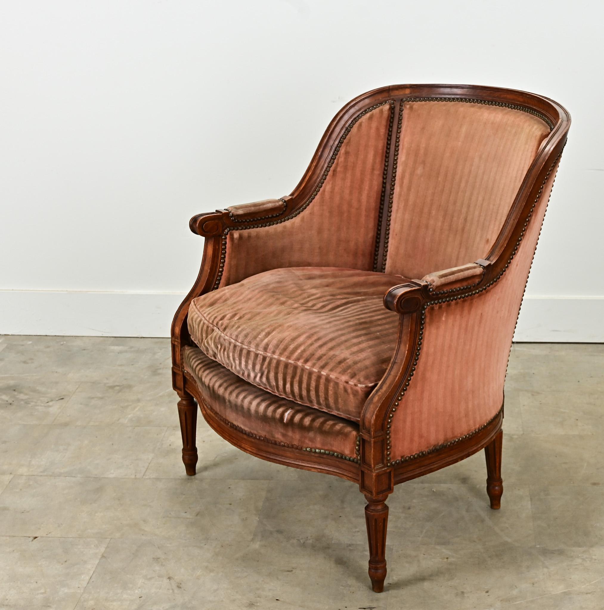 Une bergère unique de style Louis XVI datant de la fin des années 1800. Cette bergère à dossier en tonneau est fabriquée en noyer massif sculpté. Il est recouvert d'un velours rayé usé avec une garniture de tête de clou en laiton et un coussin