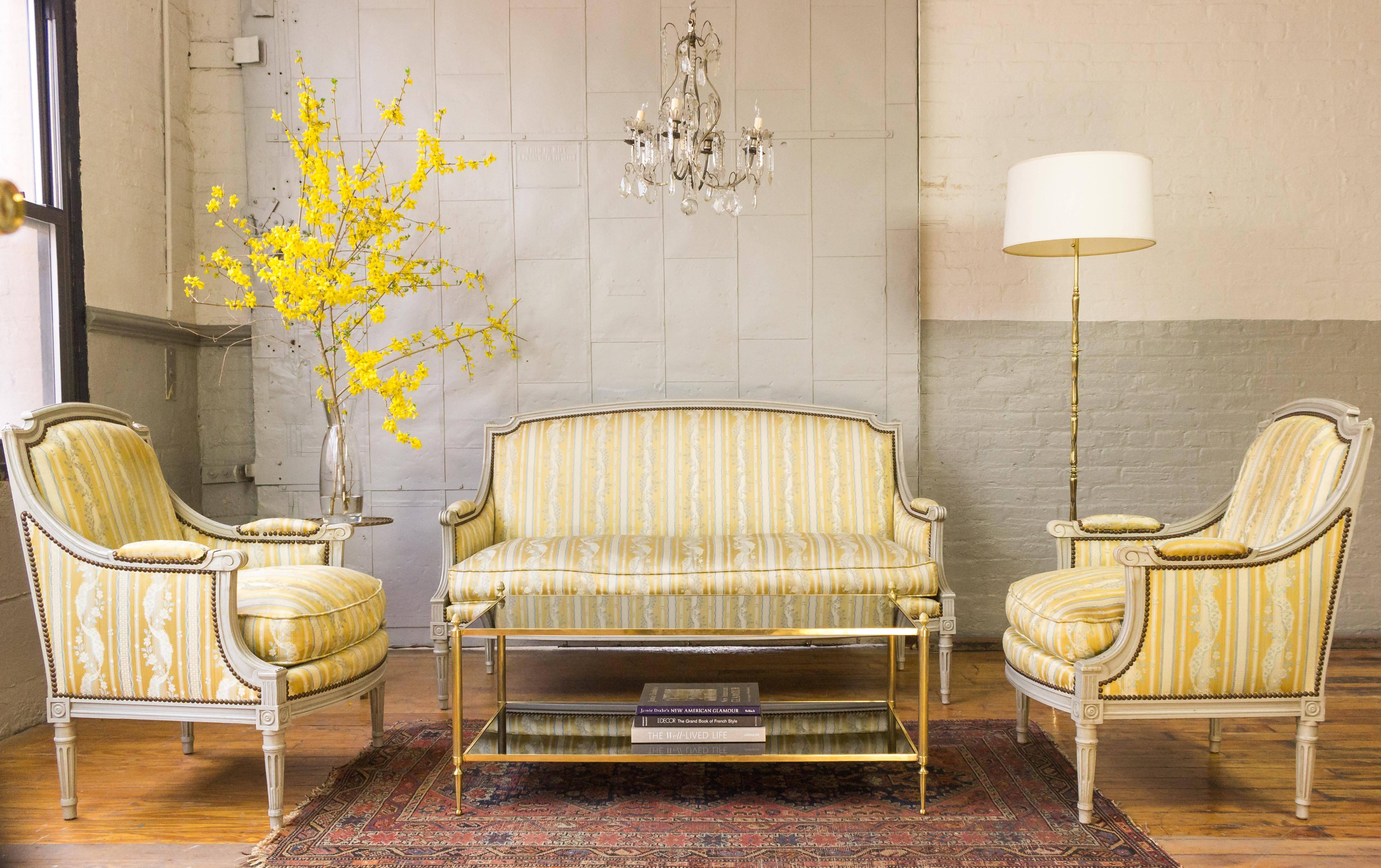French Louis XVI style sofa with a carved wooden frame with grey patina, upholstered in a striped silk fabric. The fabric is striped in white, yellow and blue with a floral design. This sofa has decorative nailheads and a loose down cushion seat. It