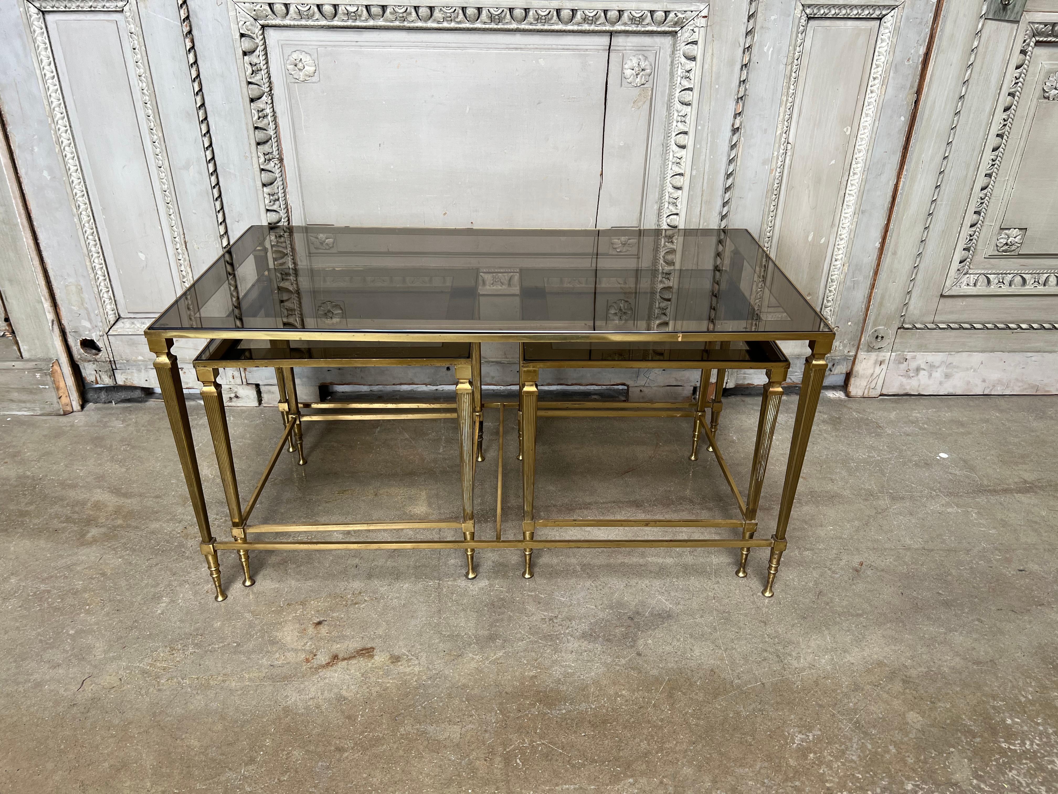 A French mid century three piece brass and glass cocktail-coffee table set with mirrored band on the smoked glass. This charming suite of tables is very useful with the two smaller tables tucking under the larger one to be pulled out when needed.