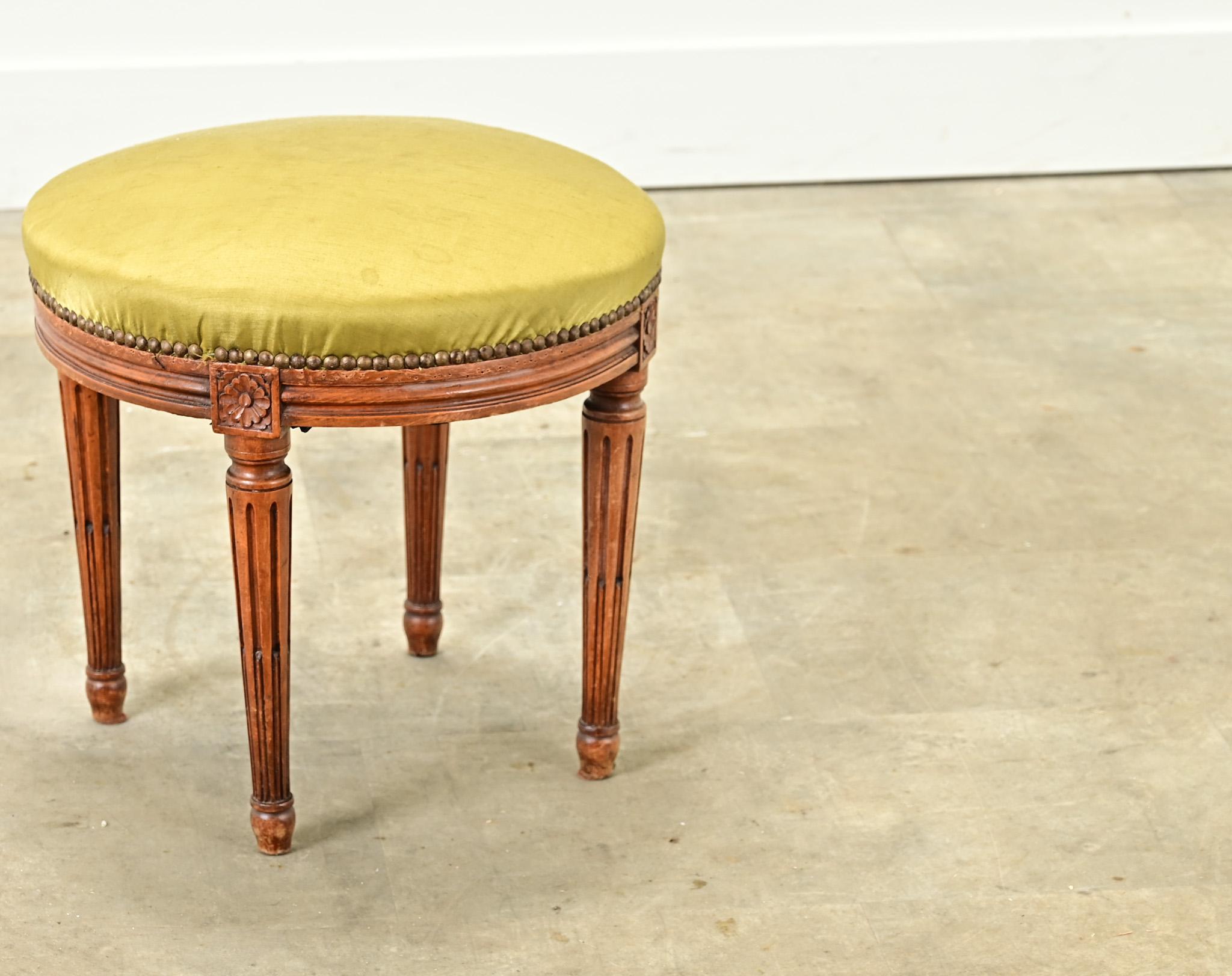 A petite French Louis XVI style stool with worn silk upholstery. The frame has classic carved rosettes with fluted and tapered legs. Be sure to view the detailed images to see the current condition.
