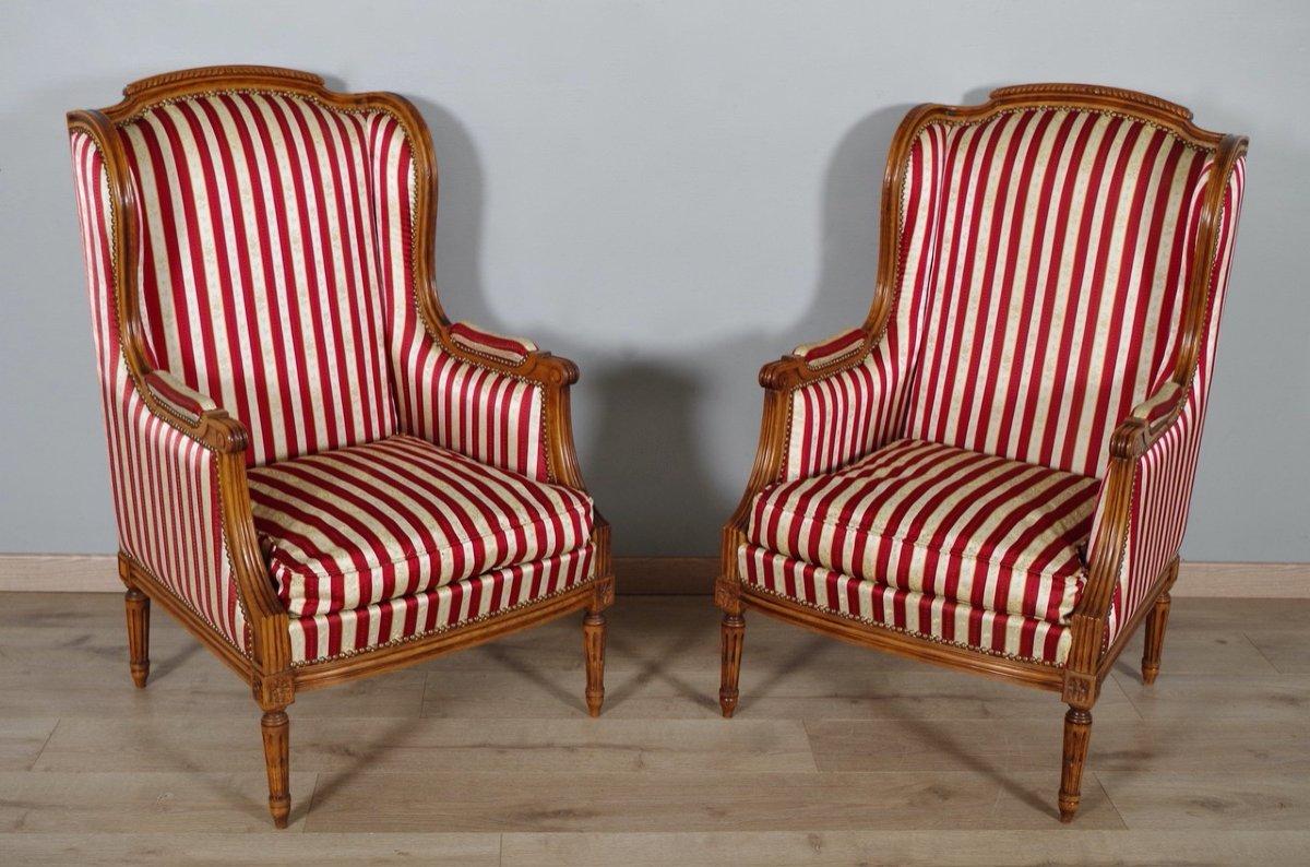 A pair of French Louis XVI style walnut bergères chairs from the 20th century with carved fluted legs and striped upholstery. Immerse yourself in the opulence of French design with this pair of Louis XVI style walnut bergère chairs, a 20th-century