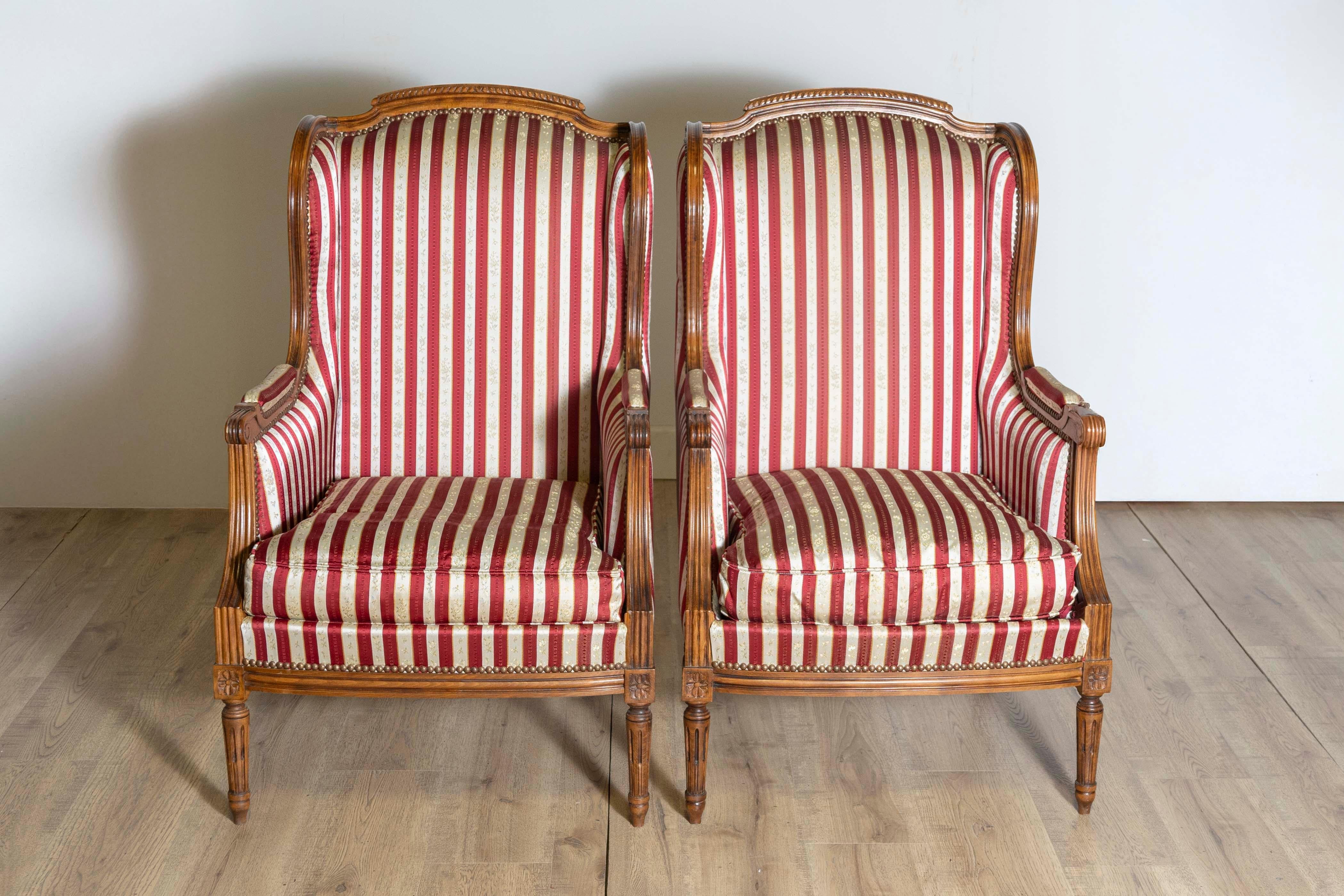 A pair of French Louis XVI style walnut bergères chairs from the 20th century with carved fluted legs and striped upholstery. Immerse yourself in the opulence of French design with this pair of Louis XVI style walnut bergère chairs, a 20th-century