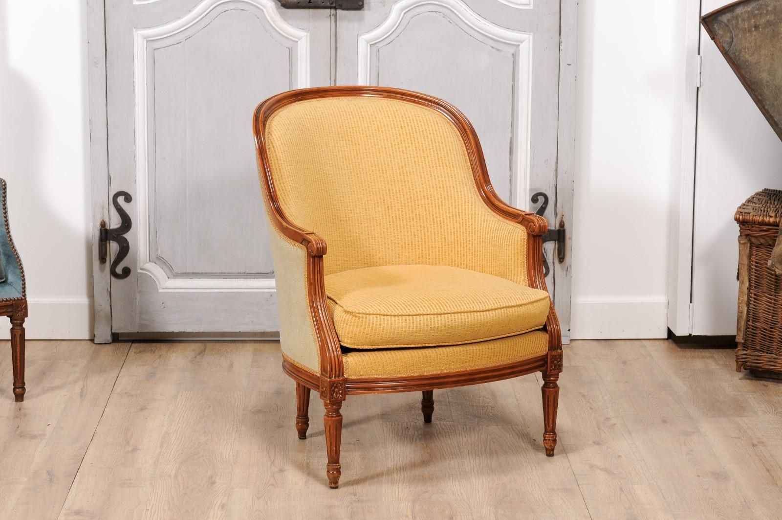 A pair of French Louis XVI style walnut bergères chairs from the 20th century with wraparound backs, scrolling knuckles on the arms, fluted legs and carved rosettes. Experience the grace and elegance of French craftsmanship with this exquisite pair