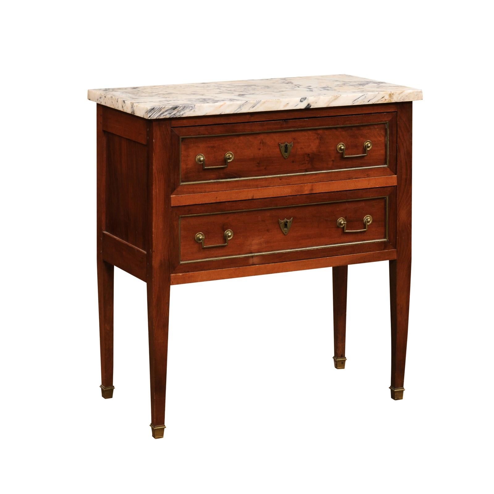 A French Louis XVI style walnut commode from the 19th century, with marble top, two drawers, tapered legs and brass hardware. Created in France during the 19th century, this walnut commode features a rectangular white and cream variegated marble top