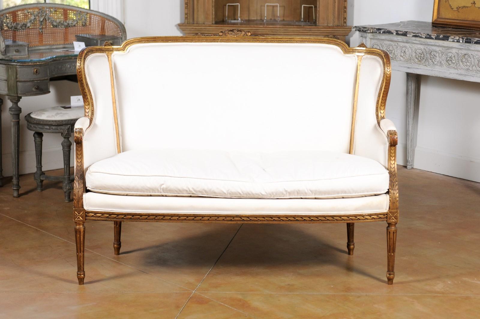 A French Louis XVI style settee from the early 19th century, with original gilding and new upholstery. Born in France during the early years of the 19th century, this settee features a wingback topped with a carved ribbon and accented with bead and