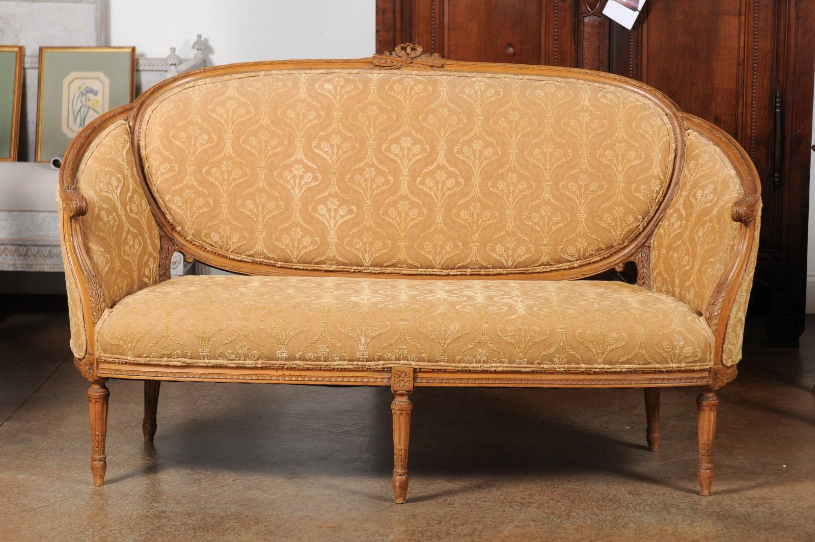 A French Louis XVI style 'canapé en corbeille' from the 19th century, with carved crest, fluted legs and upholstery. Created in France during the 19th century, this canapé features a wraparound back accented with ribbon-tied foliage and flute. The