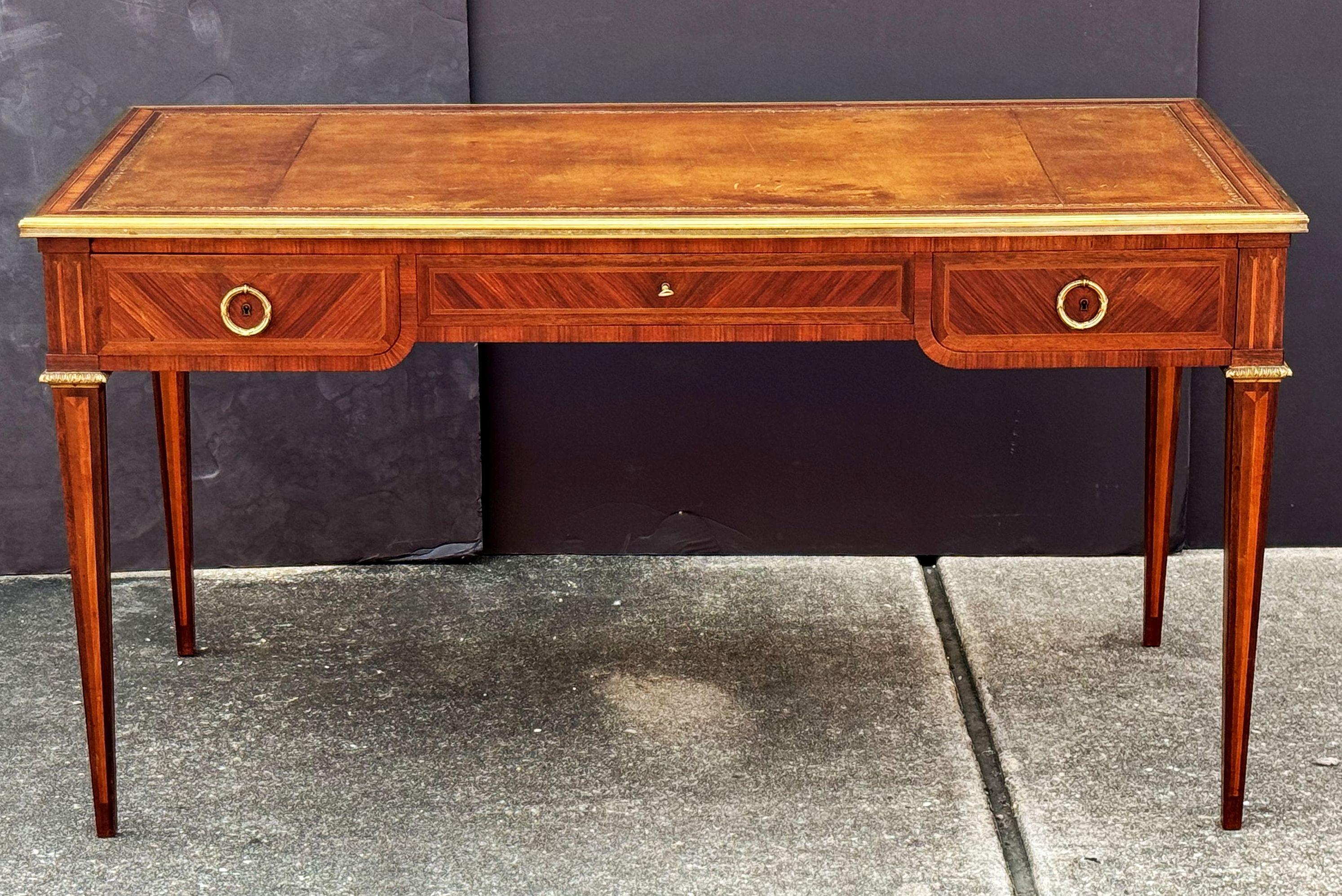 A fine French writing table or desk of ormolu mounted walnut and mahogany, in the Louis XVI style, featuring an inset embossed leather top framed with an inlaid pattern of mahogany, with brass trim around the edge, with three drawers with brass