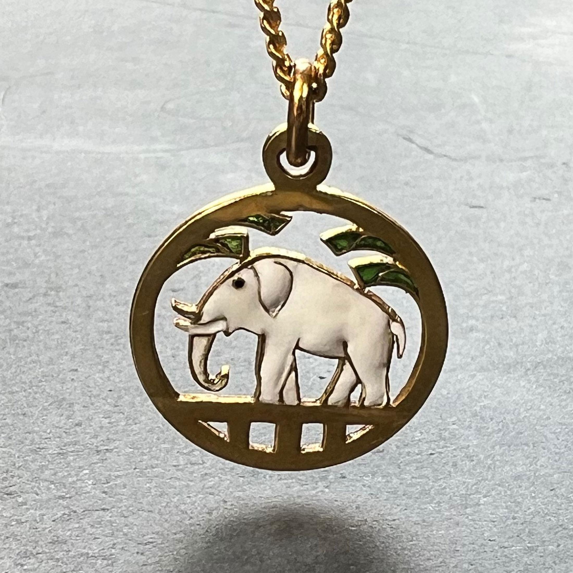 A French 18 karat (18K) yellow gold charm pendant designed as a lucky elephant with white enamel body and black enamel eye within a circular gold frame with green plique-a-jour tree branches. Stamped with the eagle's head for 18 karat gold and