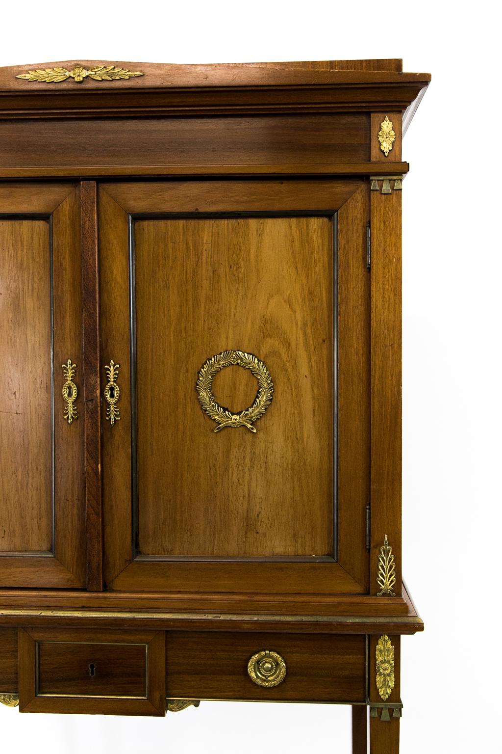 French mahogany cabinet on legs has multiple brass fittings and moldings with an architectural pediment on top. The color is somewhat faded.
  