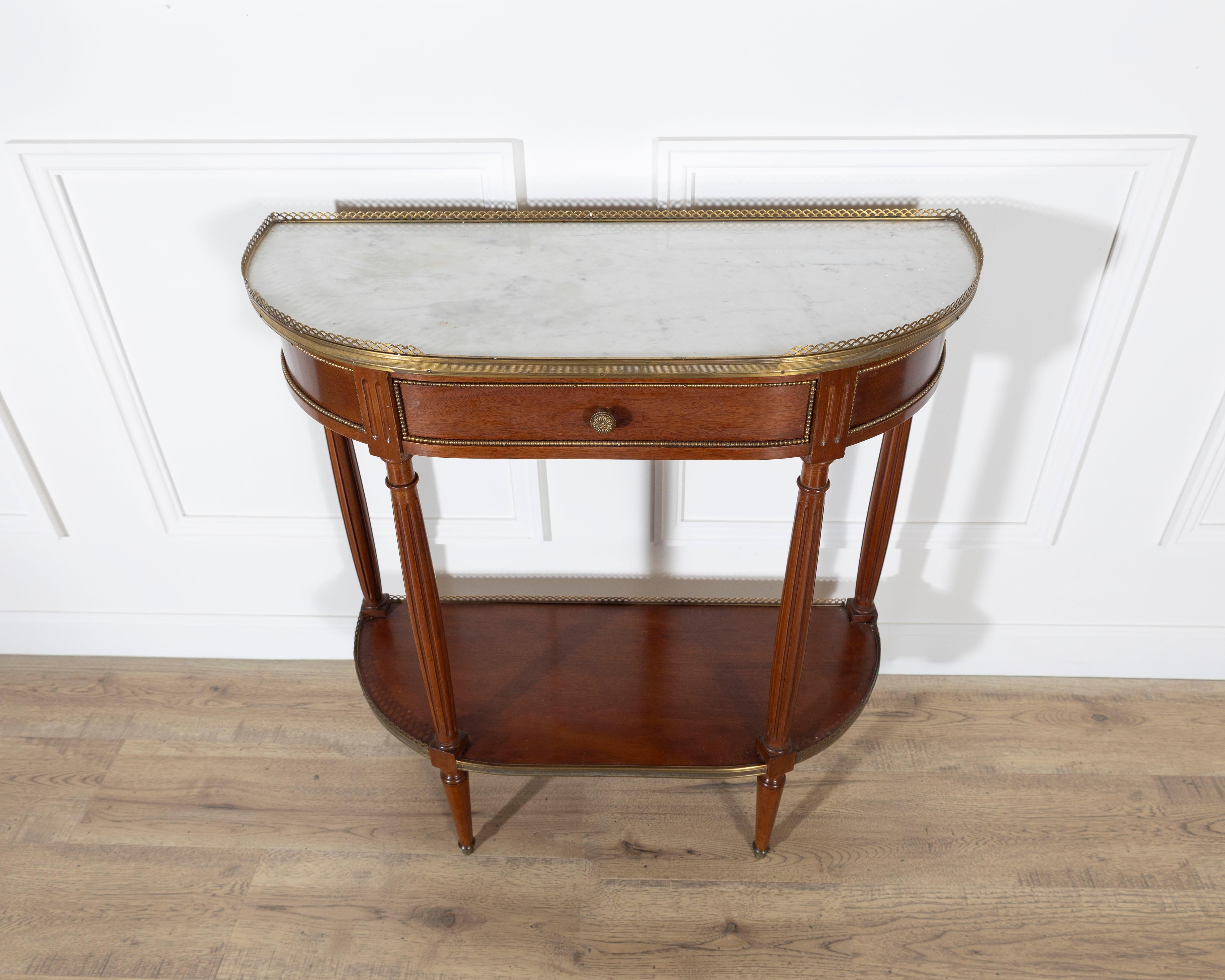 This early 20th-century French mahogany console table features a demilune form topped with a white marble surface bordered by a pierced brass gallery. The mahogany frame shows a warm, even tone and is fitted with a single central drawer set within a