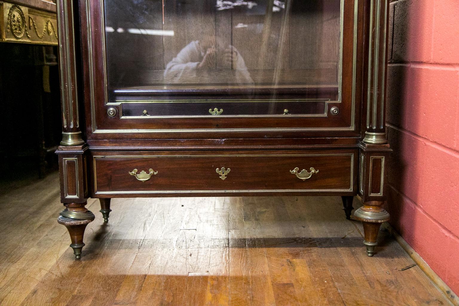 This French mahogany display cabinet or bookcase has brass trim moldings throughout with free standing columns that are fluted with inlaid brass. The interior has four adjustable shelves and a single drawer as well as an exterior drawer in the lower