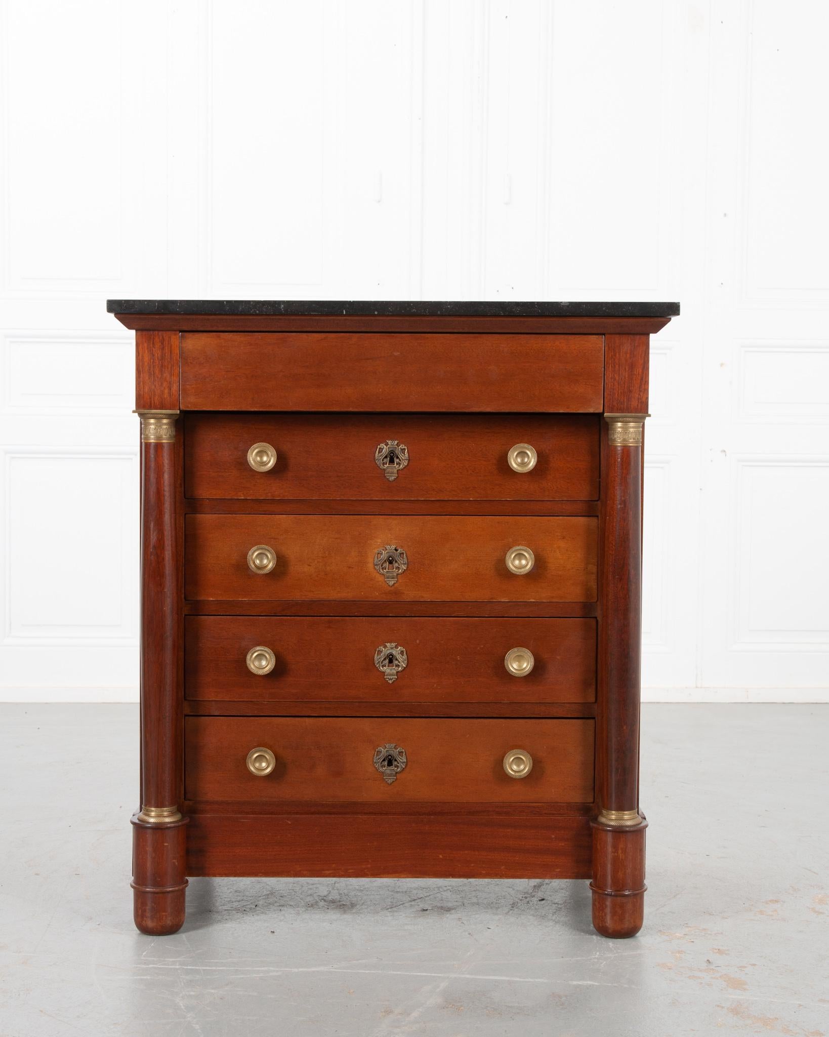 A dignified petite Empire commode from 19th century France. Unique black fossil Belgian Granite tops the mahogany base. A hidden drawer without hardware, sits flush at the top of the commode. Flanking the bank of drawers are column forms with
