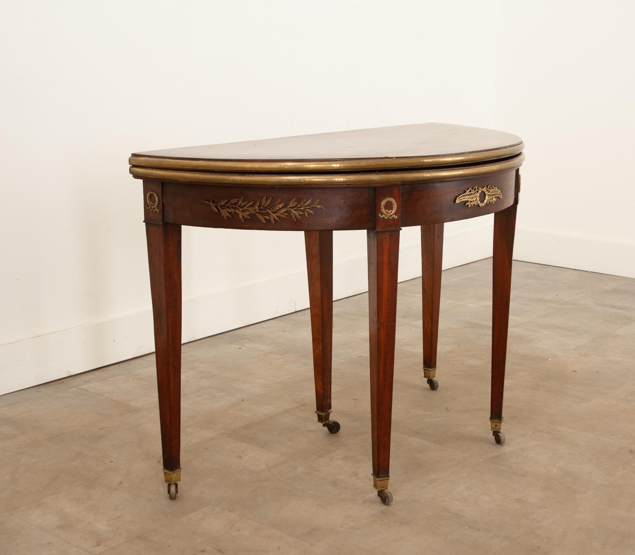 A convertible demilune console-to-game table - made in the Empire style - in 19th Century France. When closed, the table functions as a striking demilune console, bedecked in outstanding gilt brass ormolu. A drawer extends from the back of the