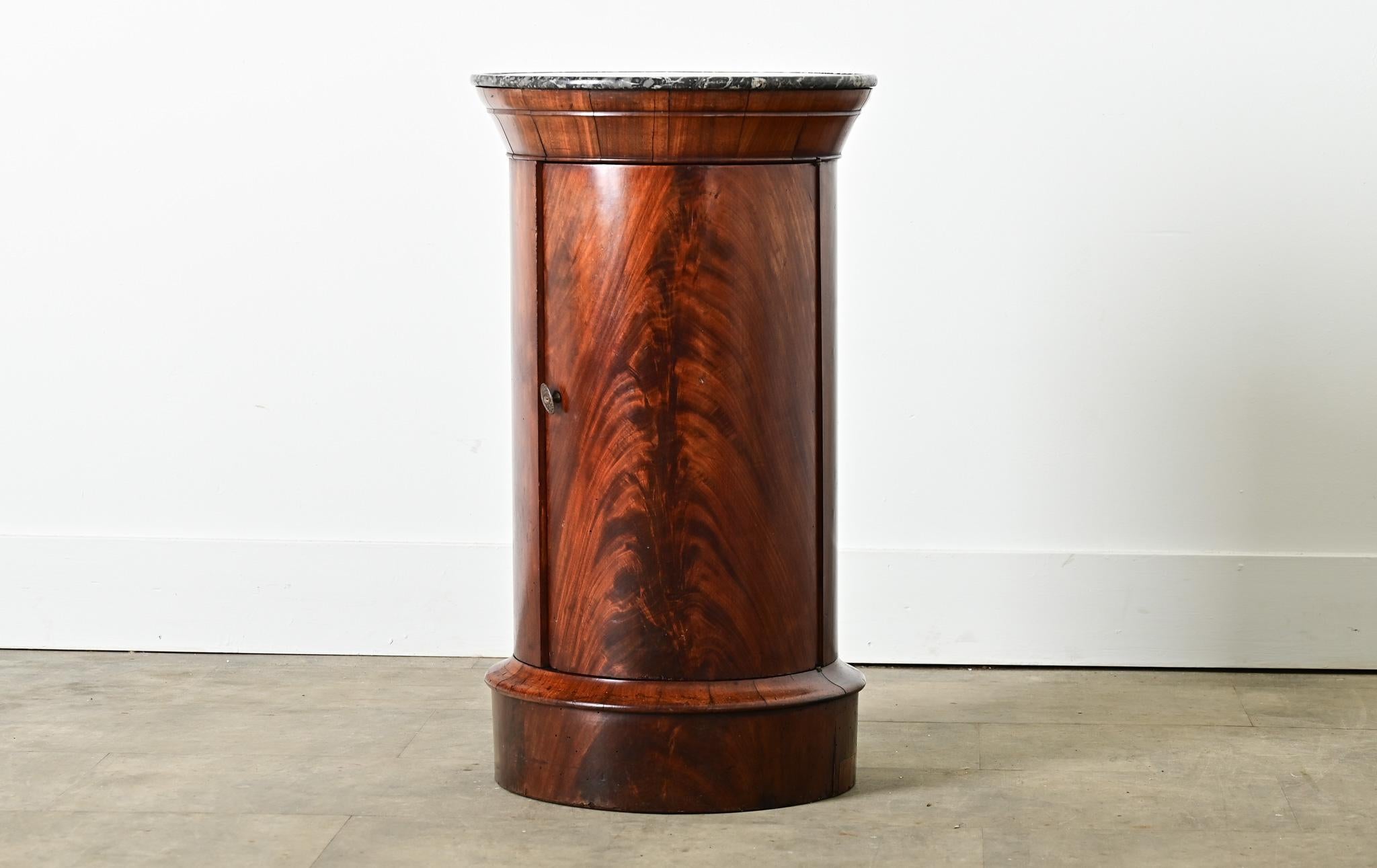 A columnar, French mahogany and marble top bedside table that offers both storage and display to fit in any room around the home. The top surface is an expressive piece of St. Anne marble with “dish top” carved to a low rim around the circumference.