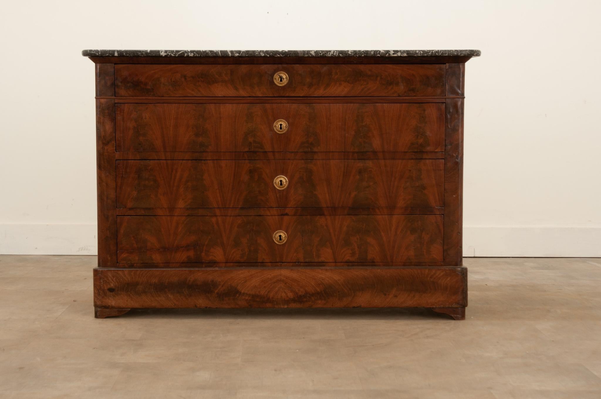 Beautifully book-matched mahogany veneer finishes this spectacular Louis Philippe style commode from 19th Century France. Numerous fossil inclusions can be found throughout the commode’s original gray marble top. The antique consists of four drawers