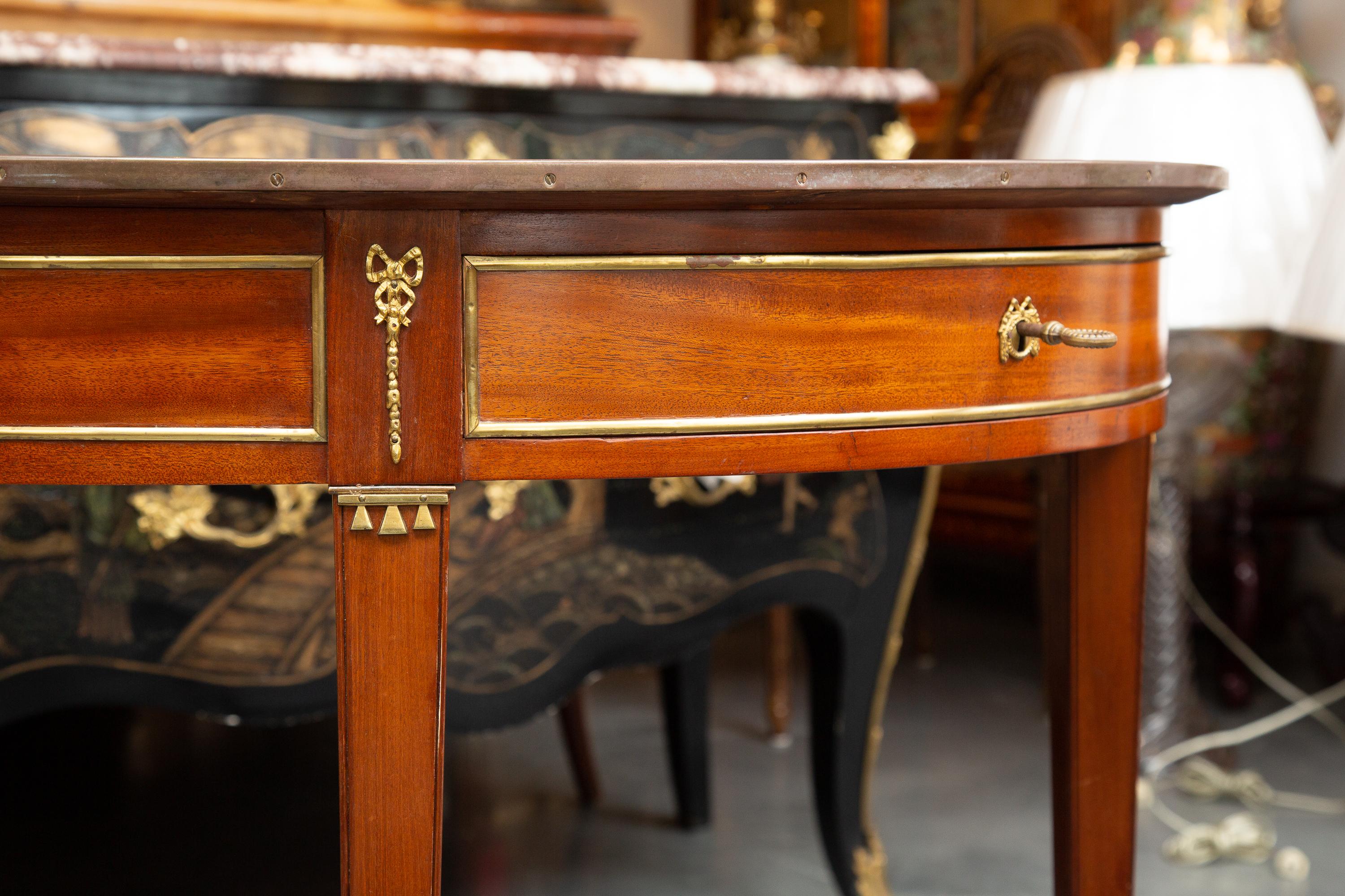 This is a soft and quiet oval mahogany Louis XVI style side table with a prominent frieze. The frieze of the table contains a drawer and raised is on straight tapered legs joined by a full stretcher. The table is trimmed with brass ornamentation
