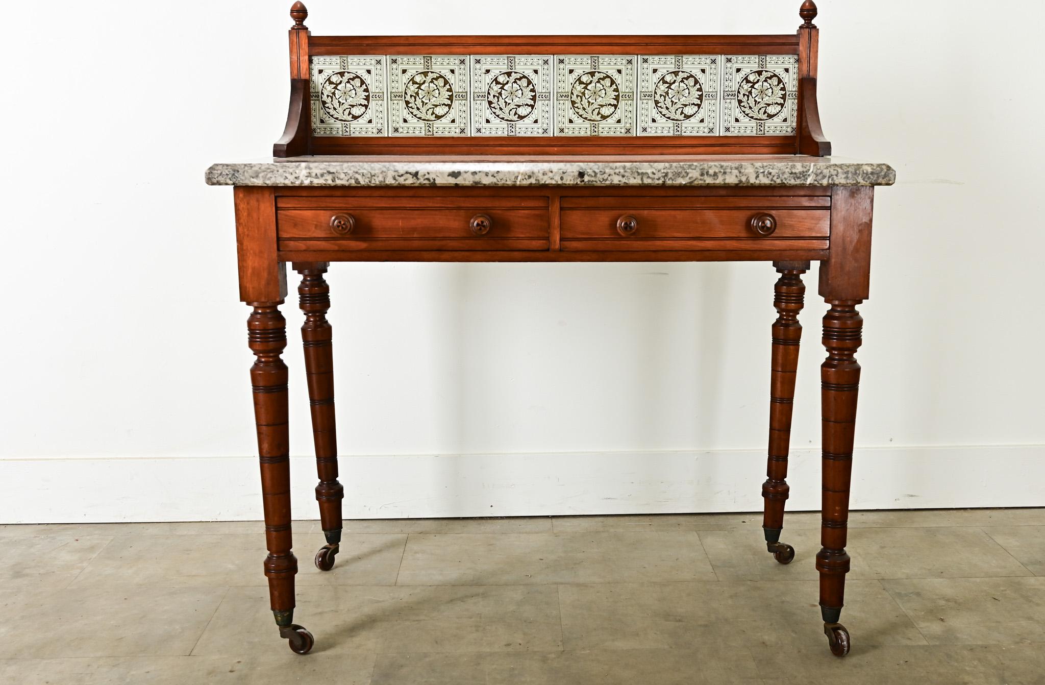 Fabulous details abound in this antique French mahogany wash stand. The backsplash, which is removable, features green and white tiles decorated with stylized, floral & foliate patterns and set within a wood frame. The speckled gray marble surface