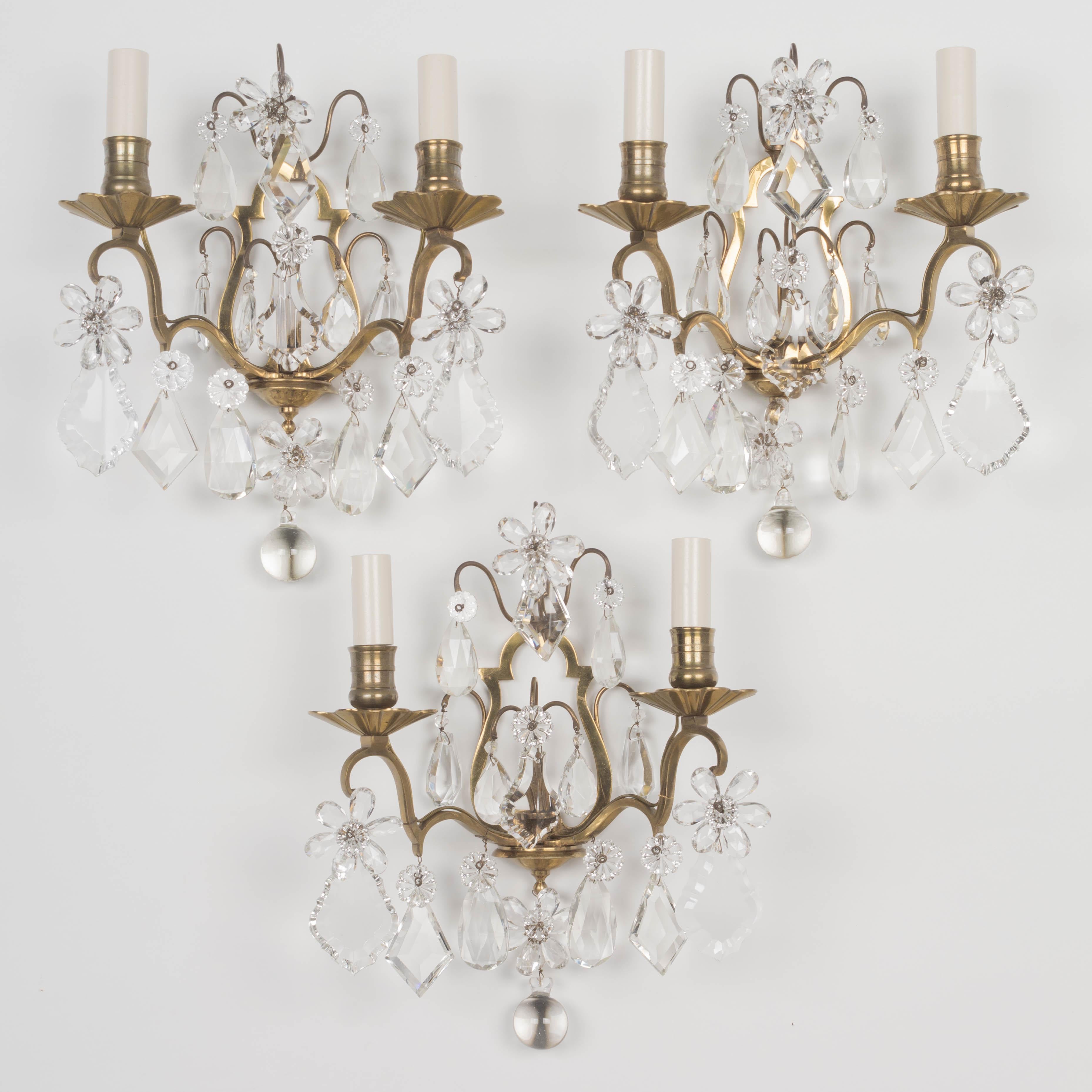 A set of three French Maison Bagues cast brass and crystal sconces. Good quality heavy casting with two scrolled candle arms and a variety of cut crystal teardrop pendalogues and faceted crystal flowers with rosettes. All original prisms. Rewired
