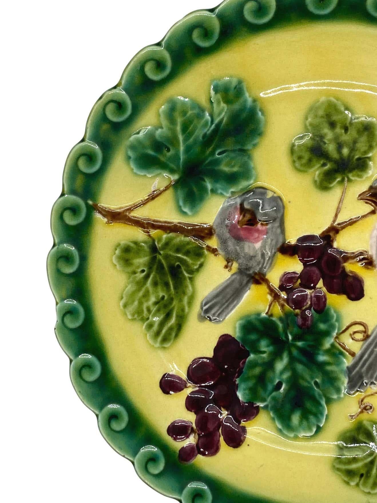 French Majolica (Barbotine) Trompe L'oeil plate, with two relief molded pink sparrows among purple grapes and green glazed leafy vines, on a yellow glazed ground, with a stylized rolling wave, scalloped and green glazed border, the reverse with