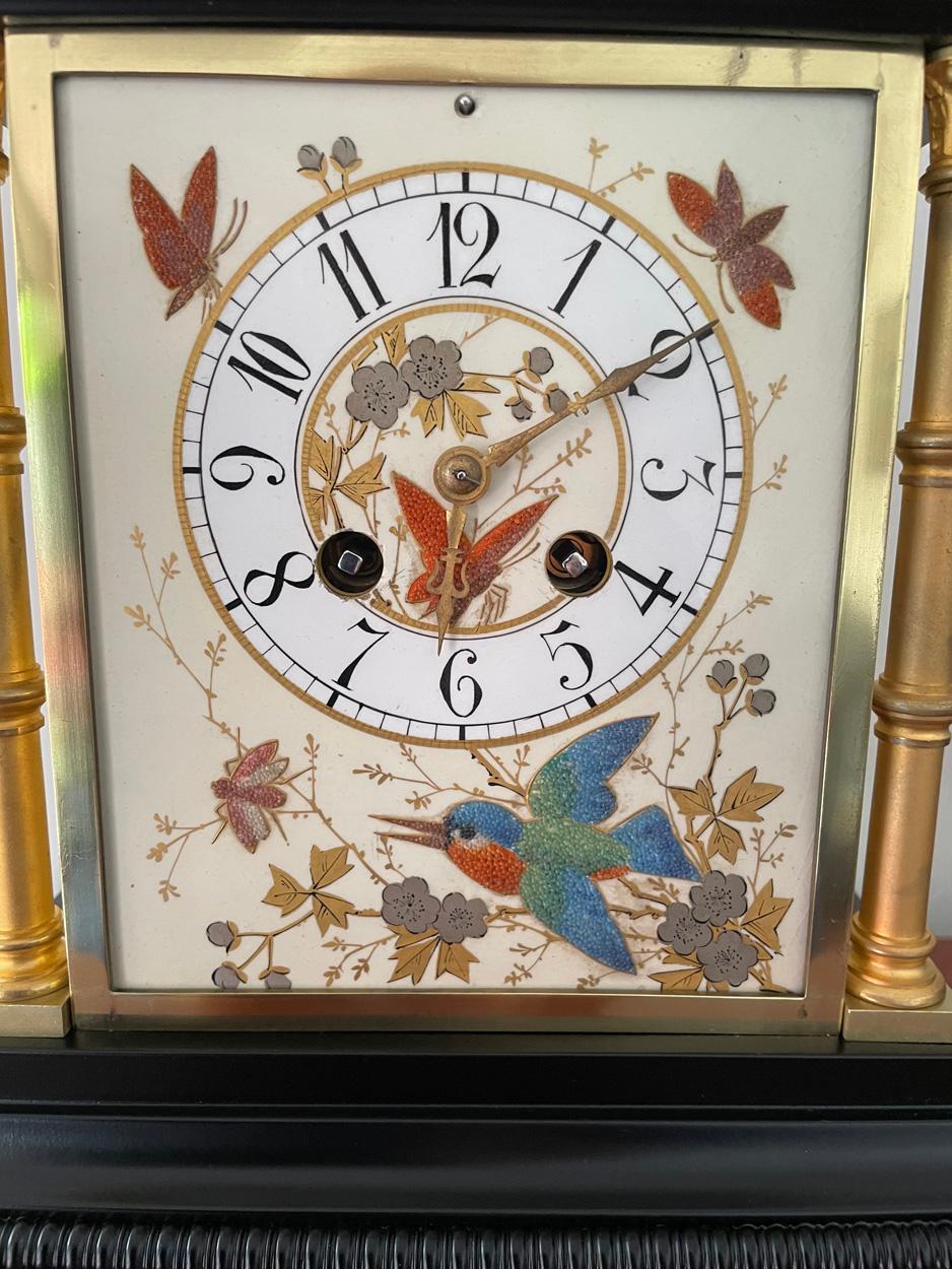 Une jolie horloge de cheminée française à boîtier en métal noir, avec des panneaux en céramique décorés d'un Kingfisher, de papillons et de feuillages, les angles étant ornés de quatre colonnes en laiton et surmontés de cinq épis de faîtage, vers