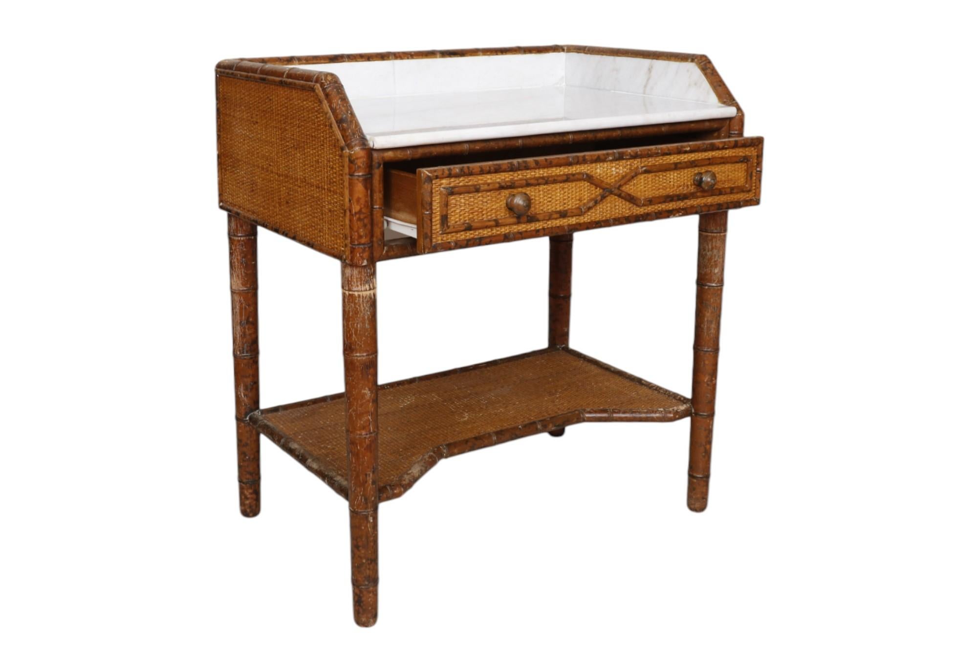 A French colonial style side table. The faux bamboo frame supports a marble table top with marble back splash and sides. A single drawer in front is decorated with strips of faux bamboo over rattan, and opens with two mushroom knob handles. The