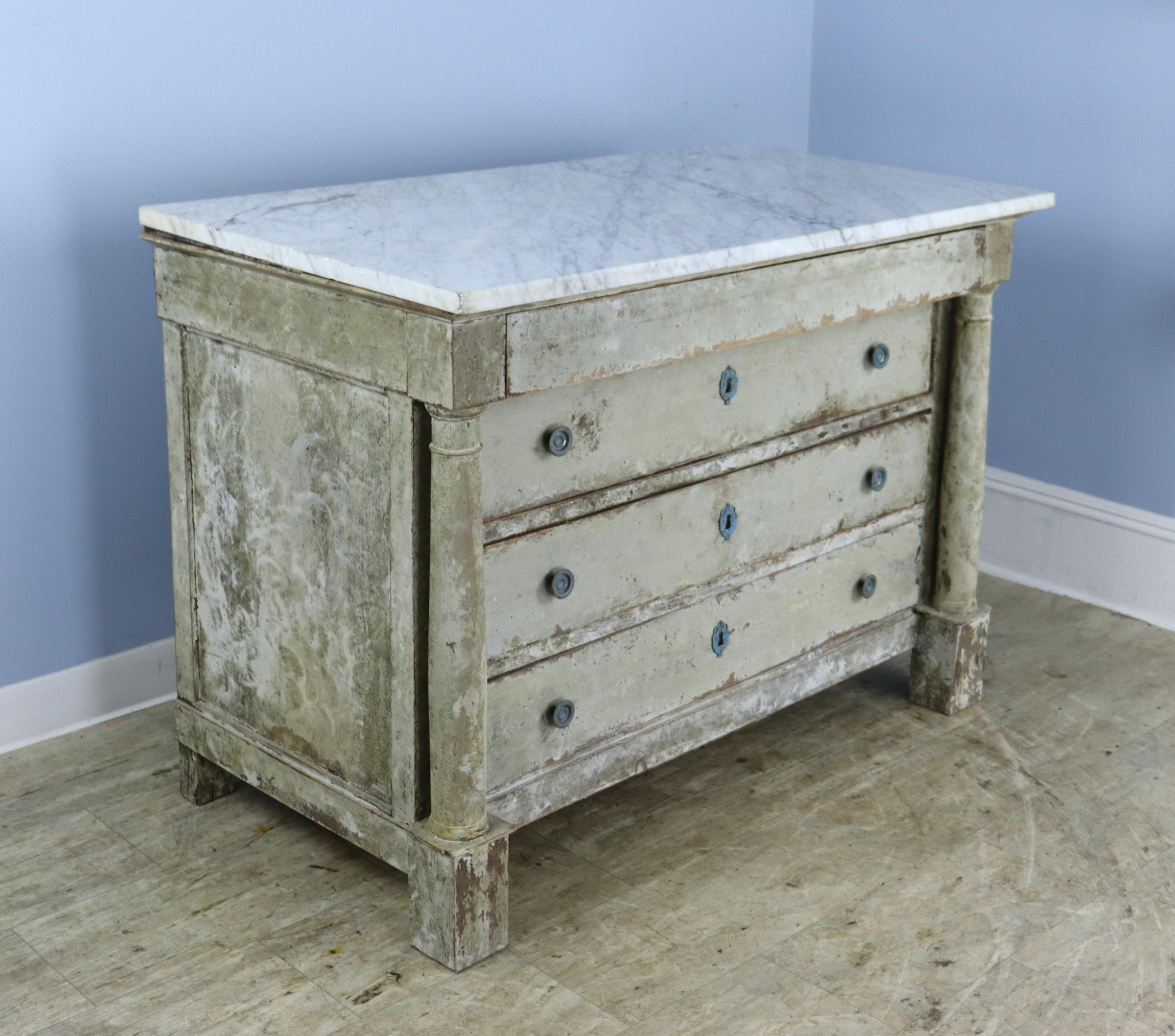 A classic Directoire commode with white marble top and column detail, newly painted in a textured faux distress style.  Four wide drawers and front block feet.  Brasses have been painted a blue green for a bright verdigris look.  Some light