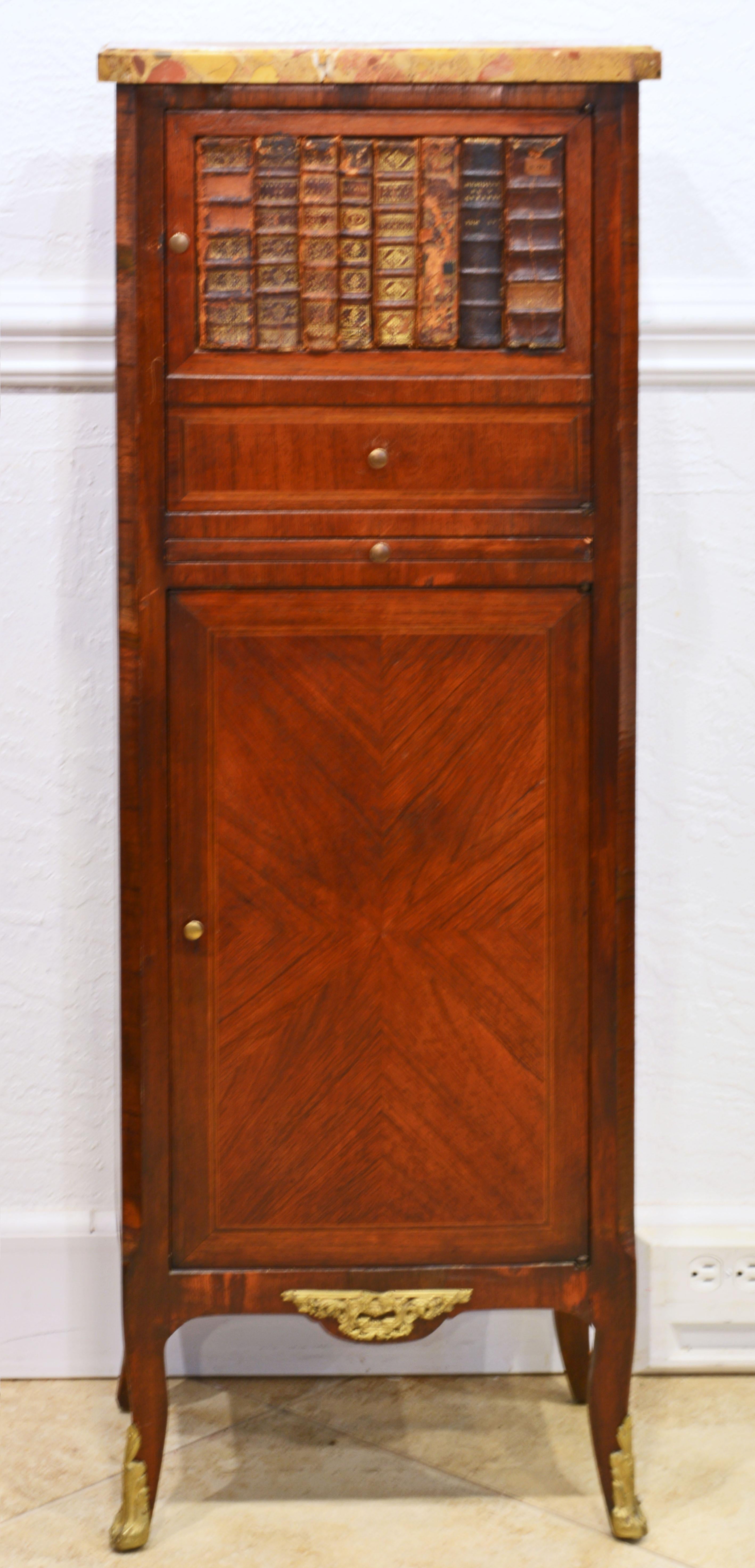 This tall but petite French cabinet, dating to the early 20th century, features a polychrome marble top above an upper cabinet with a door adorned by simulated books and drawer. Under it there is a pull out leather lined work surface and a larger