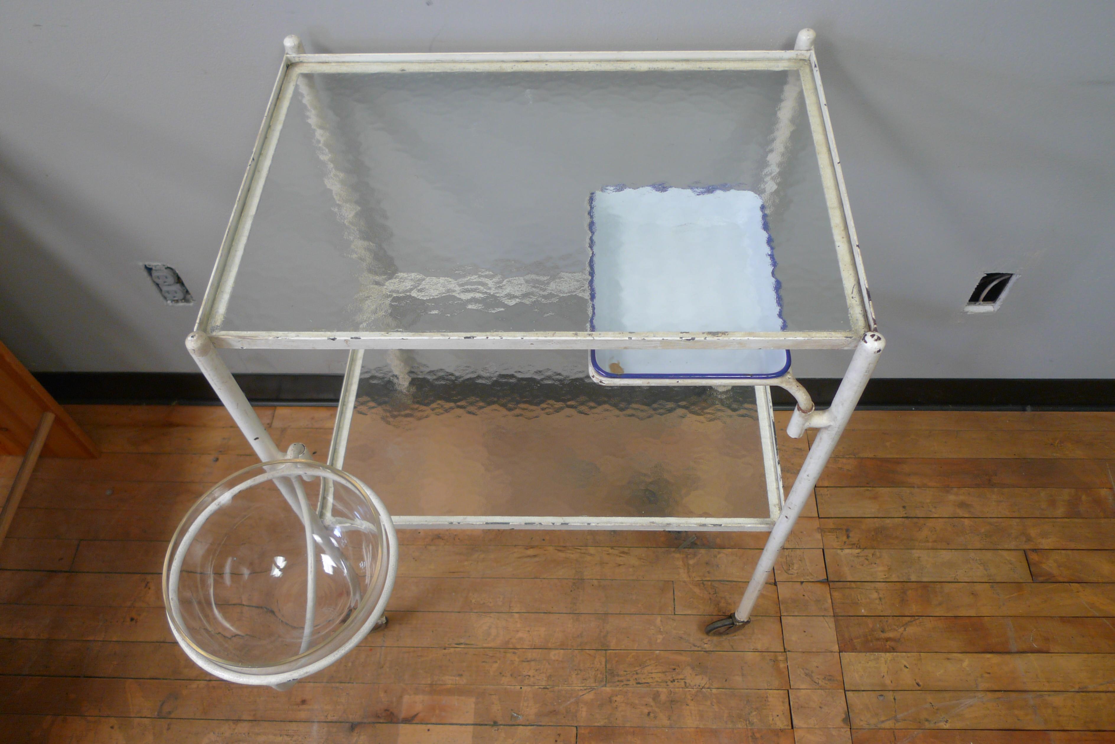 French medical cart on wheels with swing-out glass bowl and porcelain instrument tray, circa 1930s. Original wavy glass top and lower shelf have no cracks or chips. Label designates manufacturer and France as country of origin. 

For those having