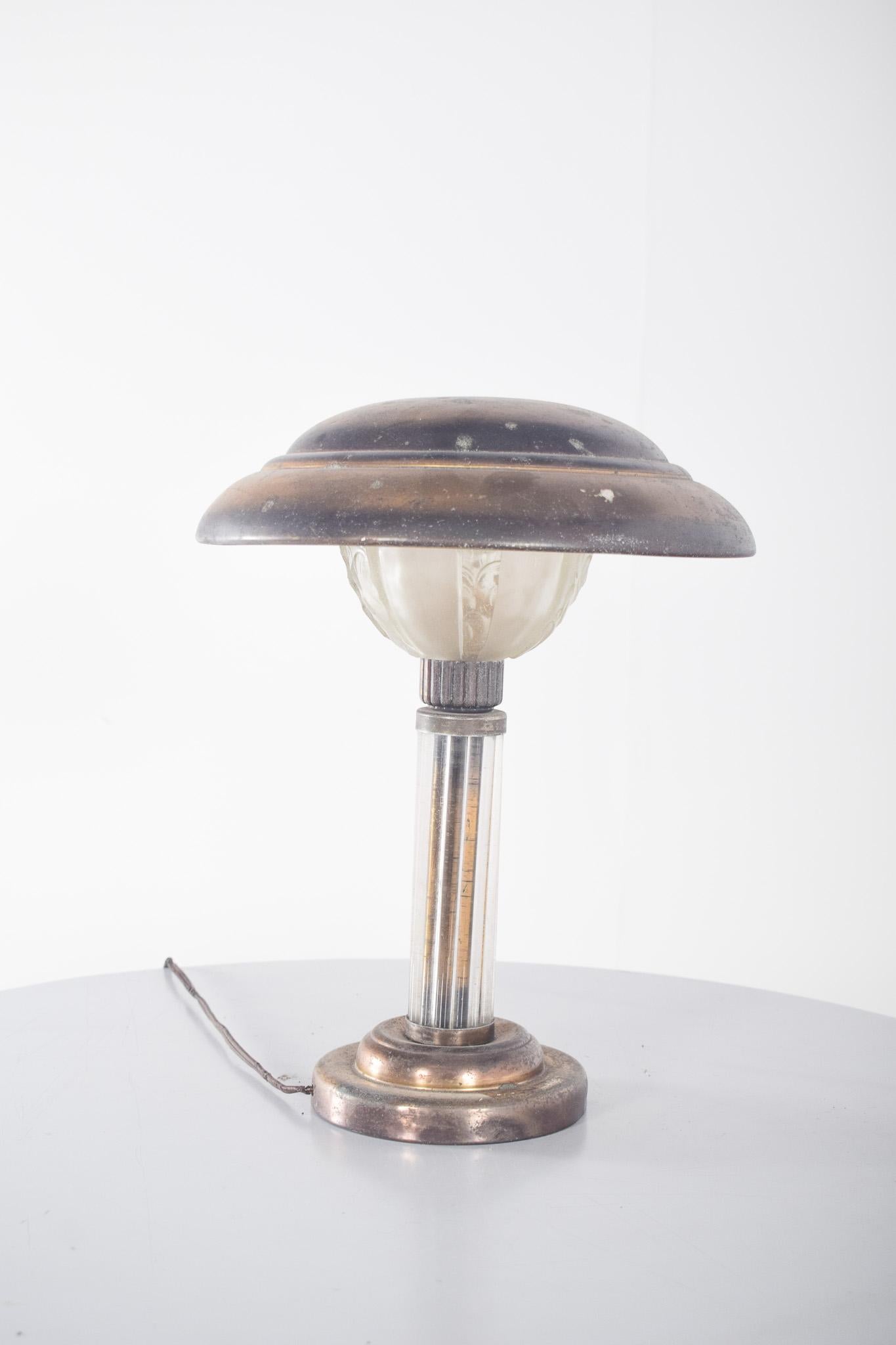 Lampada da tavolo in metallo e vetro. La robusta base in metallo ha una finitura invecchiata che le conferisce un aspetto rustico e autentico. Il gambo centrale in vetro traslucido aggiunge leggerezza alla composizione, creando un contrasto