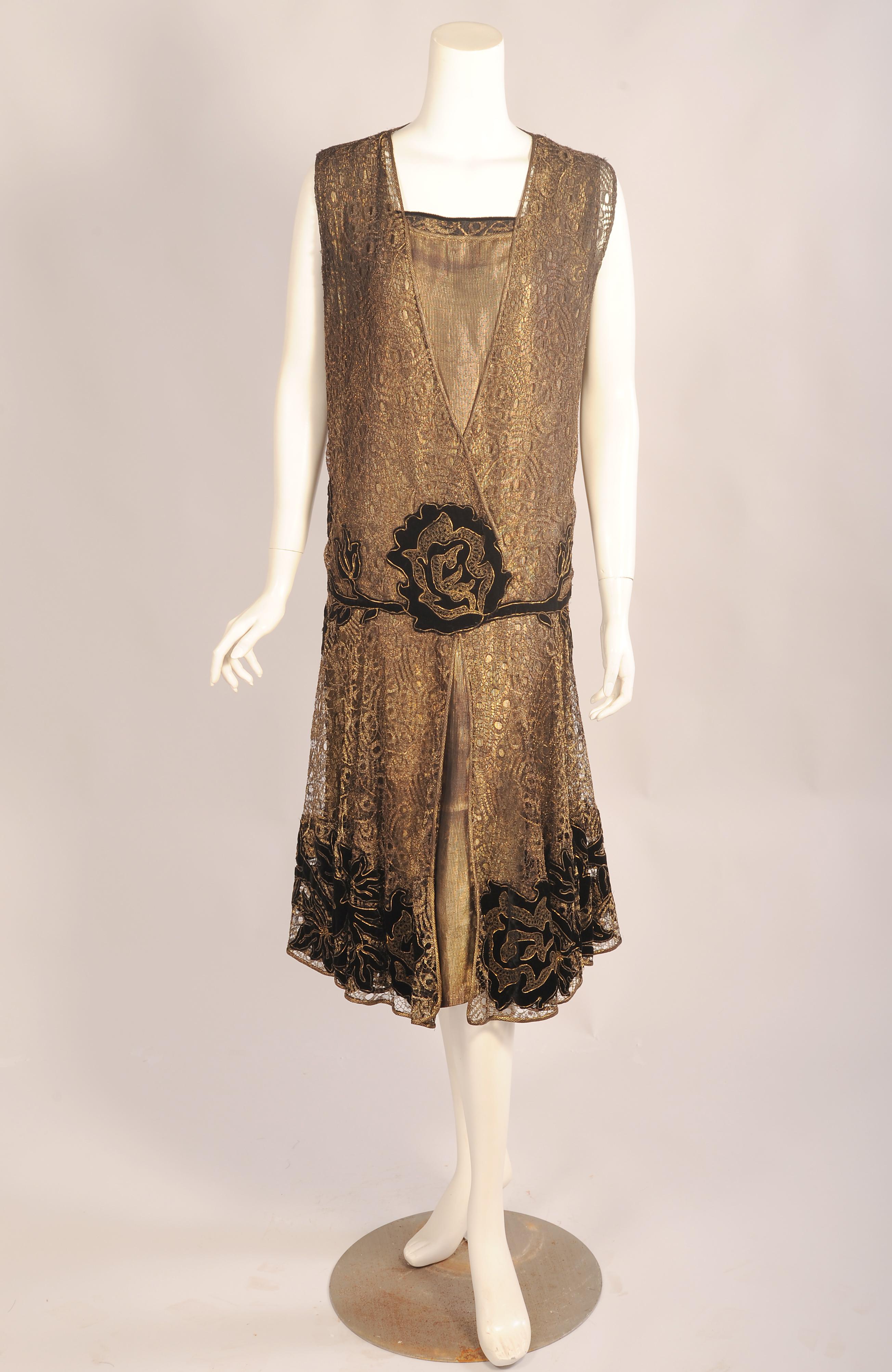 Glistening metallic gold lace is layered over a gold lame slip for this dress by Braeburn, Paris and New York. The sleeveless dress has a wrap style bodice above the dropped waist which is appliqued with black velvet blossoms outlined with gold