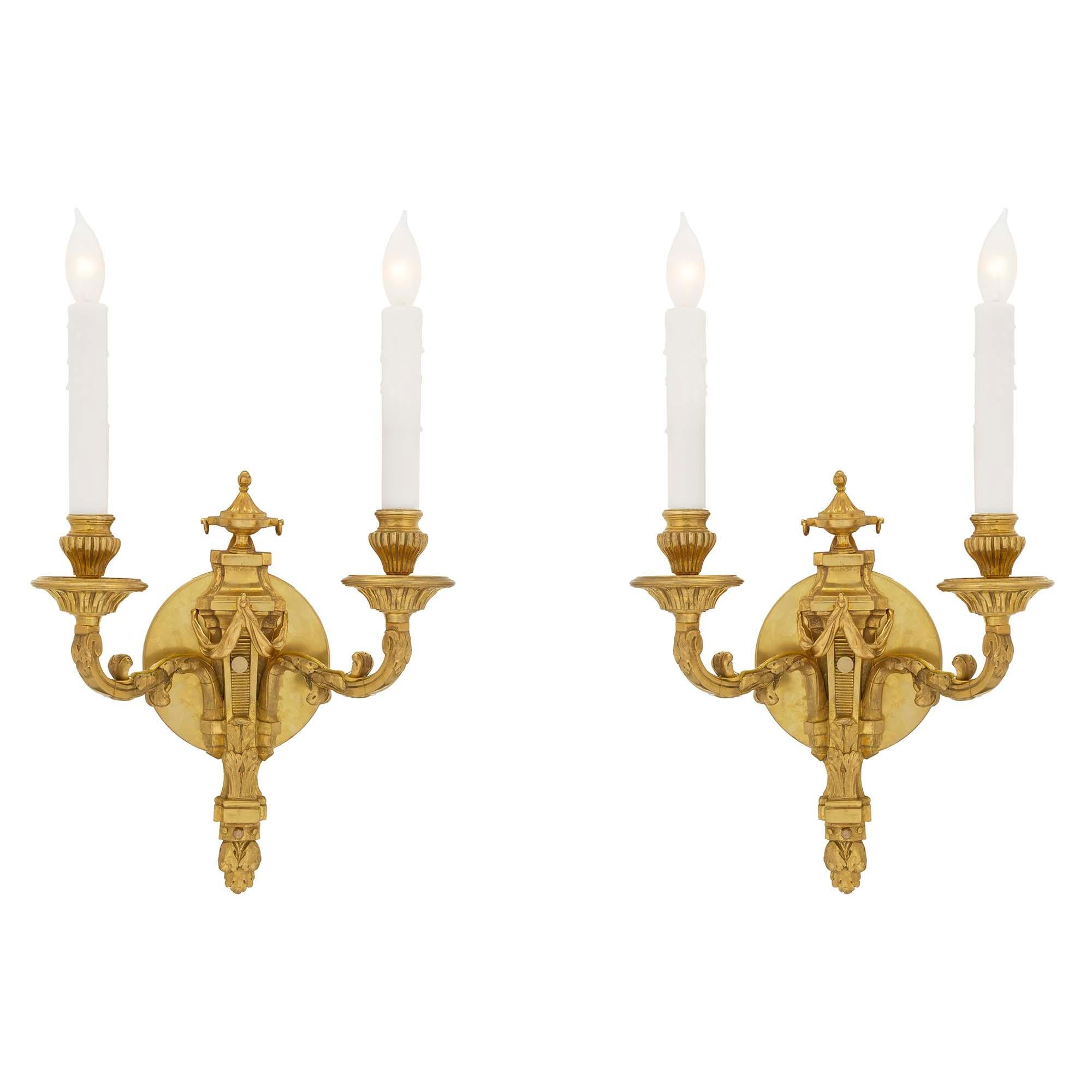 An elegant pair of French mid-19th century Louis XVI ormolu sconces. Each backplate has an urn of prosperity above a draped ribbon. Below is a recessed reeded design above a double acanthus leave with a bottom final. The two electrified ‘S’ scrolled