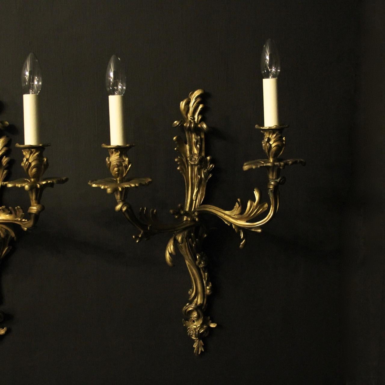 A quality French pair of gilded cast bronze twin arm electrically converted opposing antique candle wall sconces, the ornate scrolling arms with leaf bobeche drip pans and bulbous leaf candle sconces, issuing from a decoratively cast opposing leaf