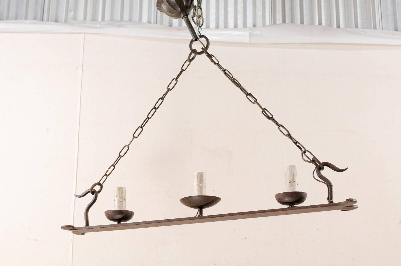 A French iron three-light vintage chandelier. This French rectangular-shaped chandelier from the mid-20th century, features a flattened, oblong forged-iron beam with decorative split-ends, topped with three cupped iron bobèches and painted candle