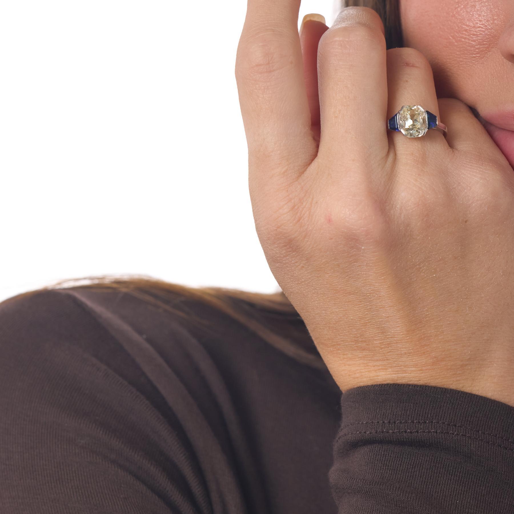 Cette bague française en platine du milieu du siècle met en valeur un captivant diamant taille ancienne dont les larges facettes et la teinte chaude créent un éclat profond au centre de la conception. Flanquée de deux saphirs de taille trapézoïdale