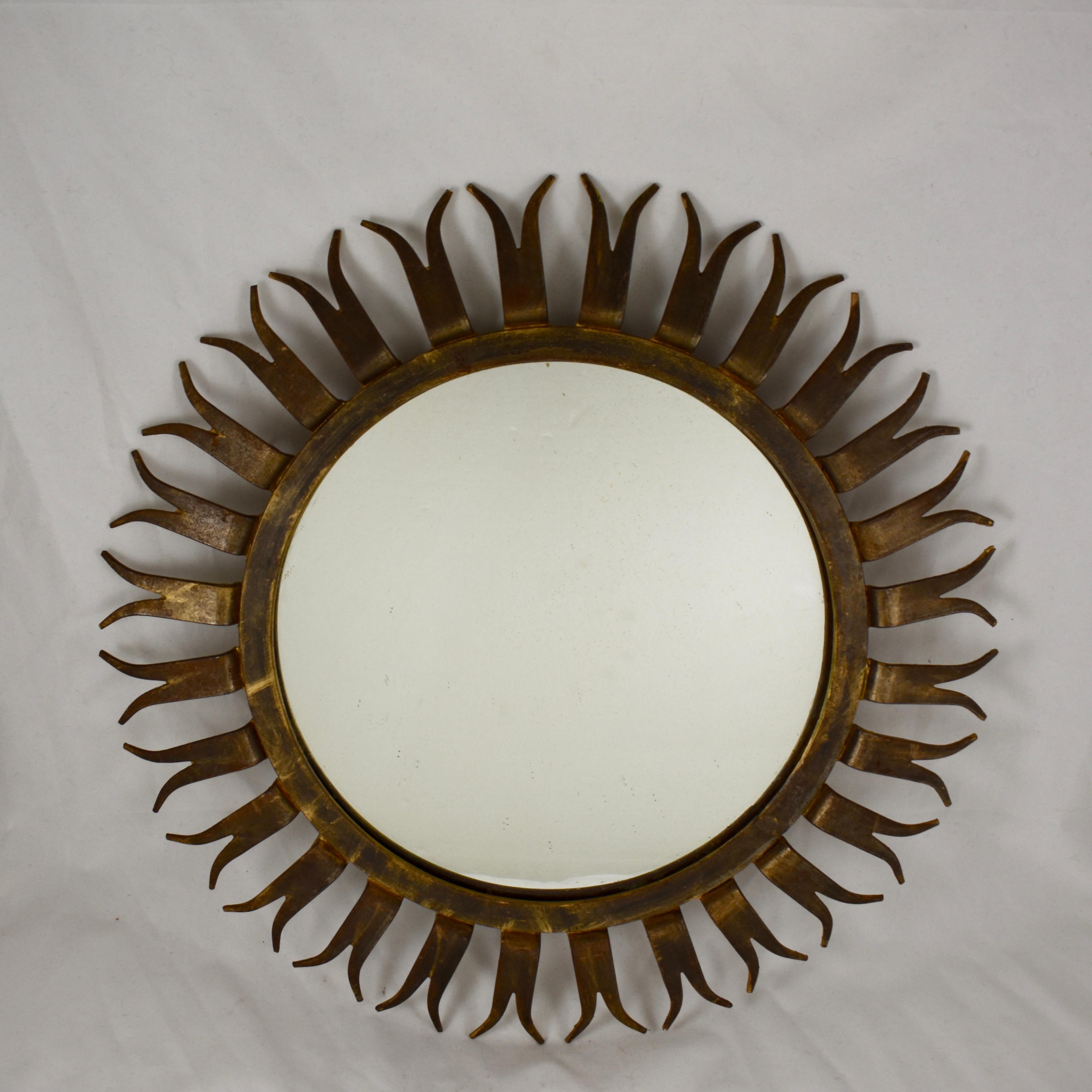 A large and heavy, midcentury French, gilded wrought iron sunburst wall mirror. Forked shaped sun rays extend from a wide bezel framing a round mirror. The rays bend slightly inward from the bezel, towards the wall when hung, adding a nice sense of