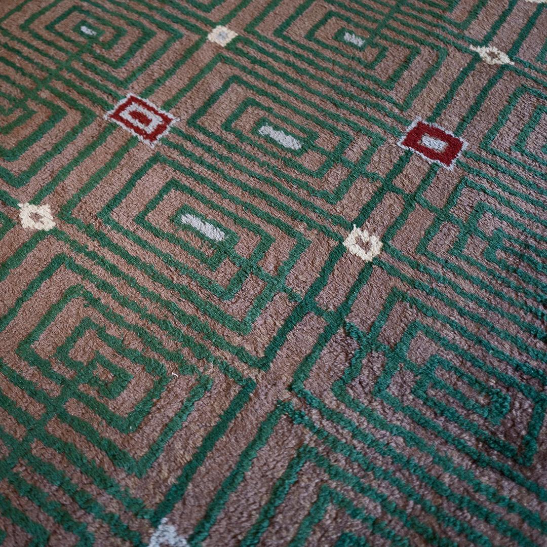 This handwoven midcentury French Deco rug, signed Maison Jules Leleu, has a brown field with a profusion of green spirals peppered with a scattering of yellow, burgundy and light blue geometric icons.