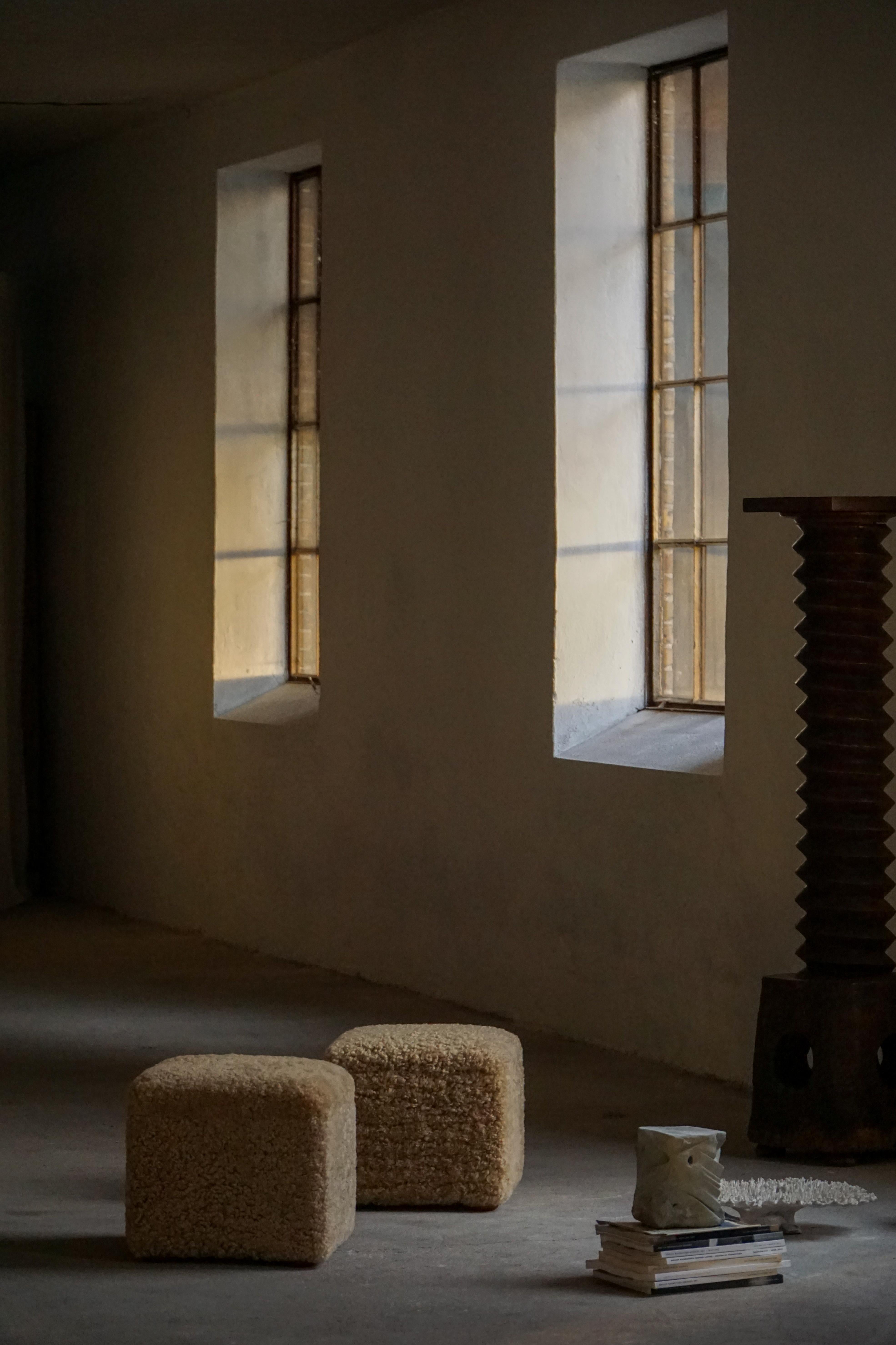 Paire de poufs cubiques modernes, nouvellement revêtus de laine d'agneau miel clair, offrant une texture douce et accueillante. Chaque pouf repose sur quatre petits pieds arrondis, créant un subtil effet de 
