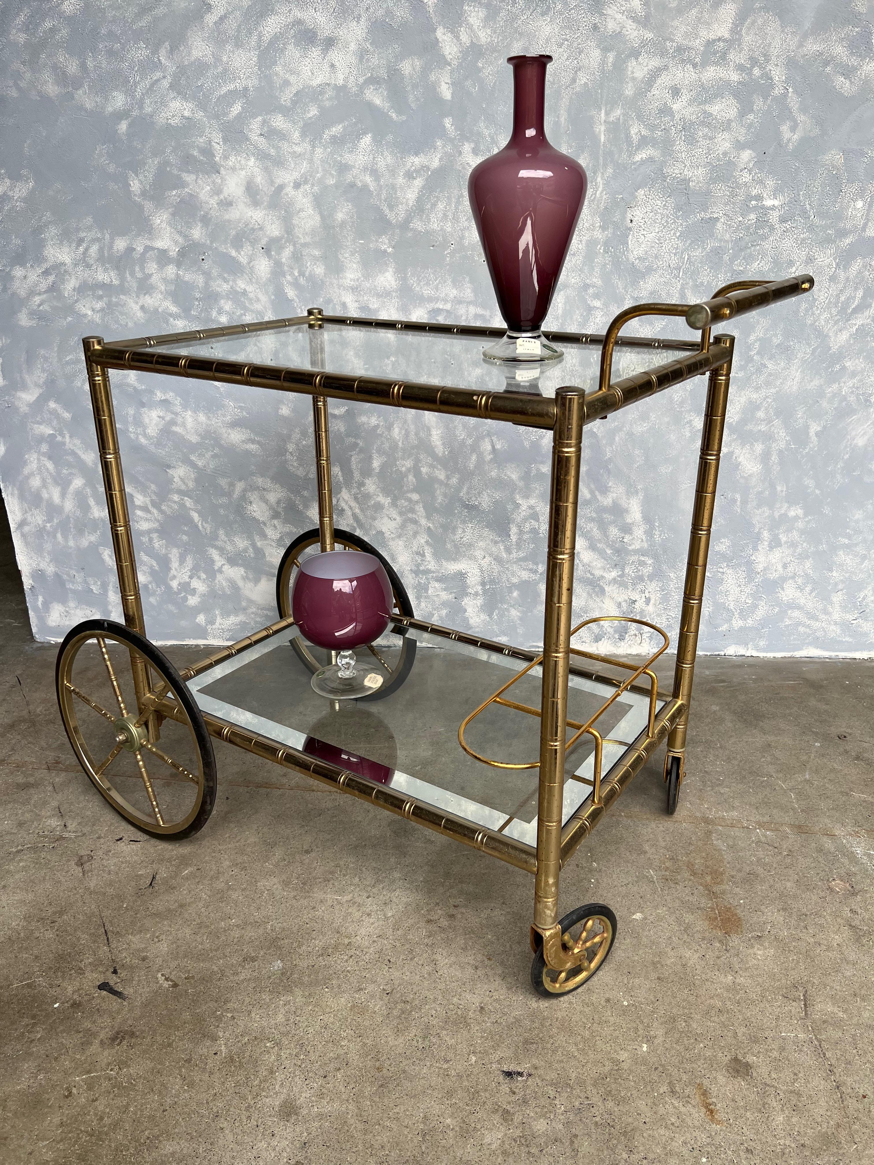An elegant French brass and glass bar cart from the 1950s. The brass frame has a stylized bamboo motif and is supported on large rubberized wheels on the front and smaller ones on the back with a handle to allow for easy moving. The original glass