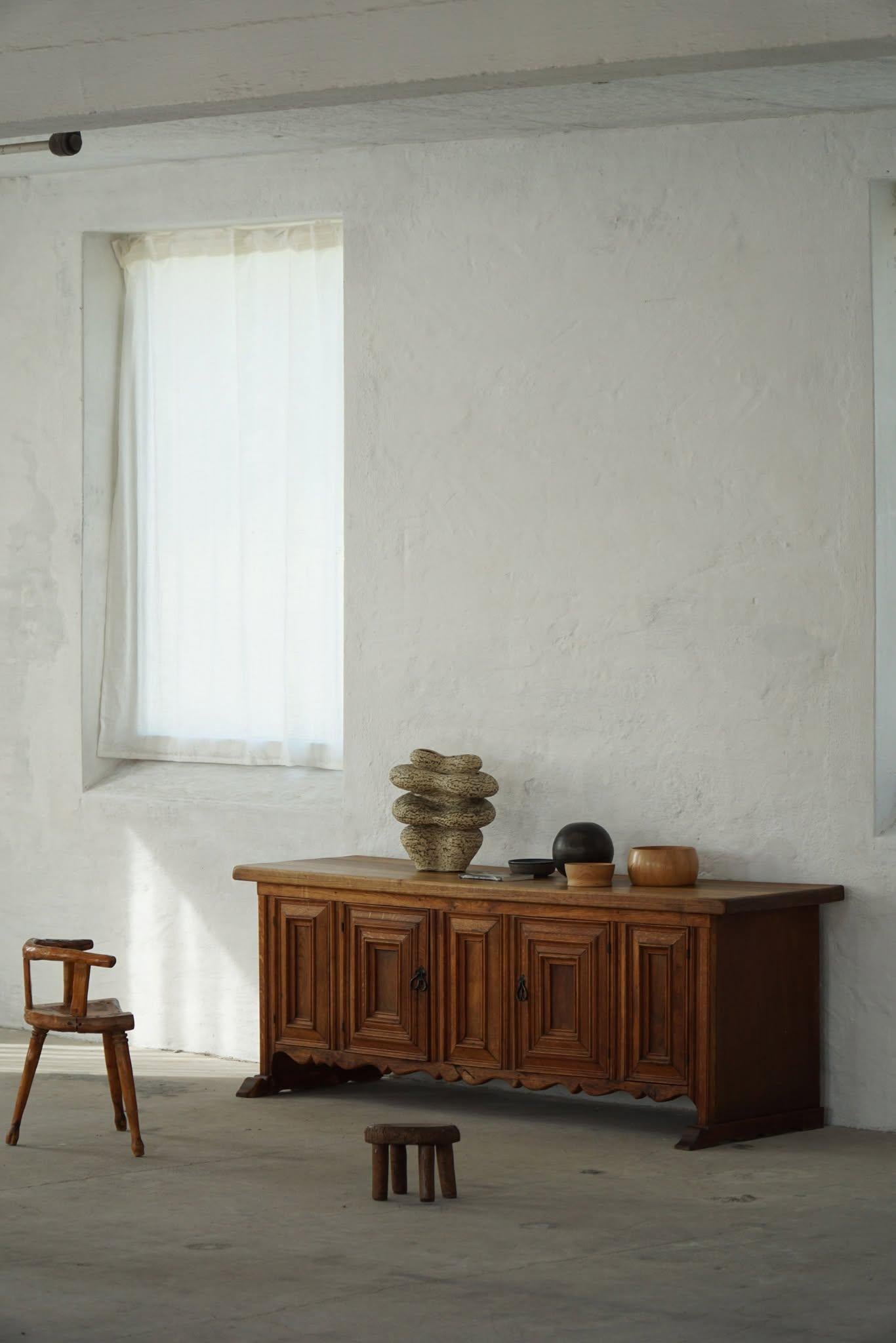 Una sostanziosa credenza bassa realizzata in rovere massiccio negli anni '40-'50, progettata in stile revival barocco. La facciata è caratterizzata da quattro porte a pannelli con maniglie in ferro forgiato, che si aprono per rivelare scaffali