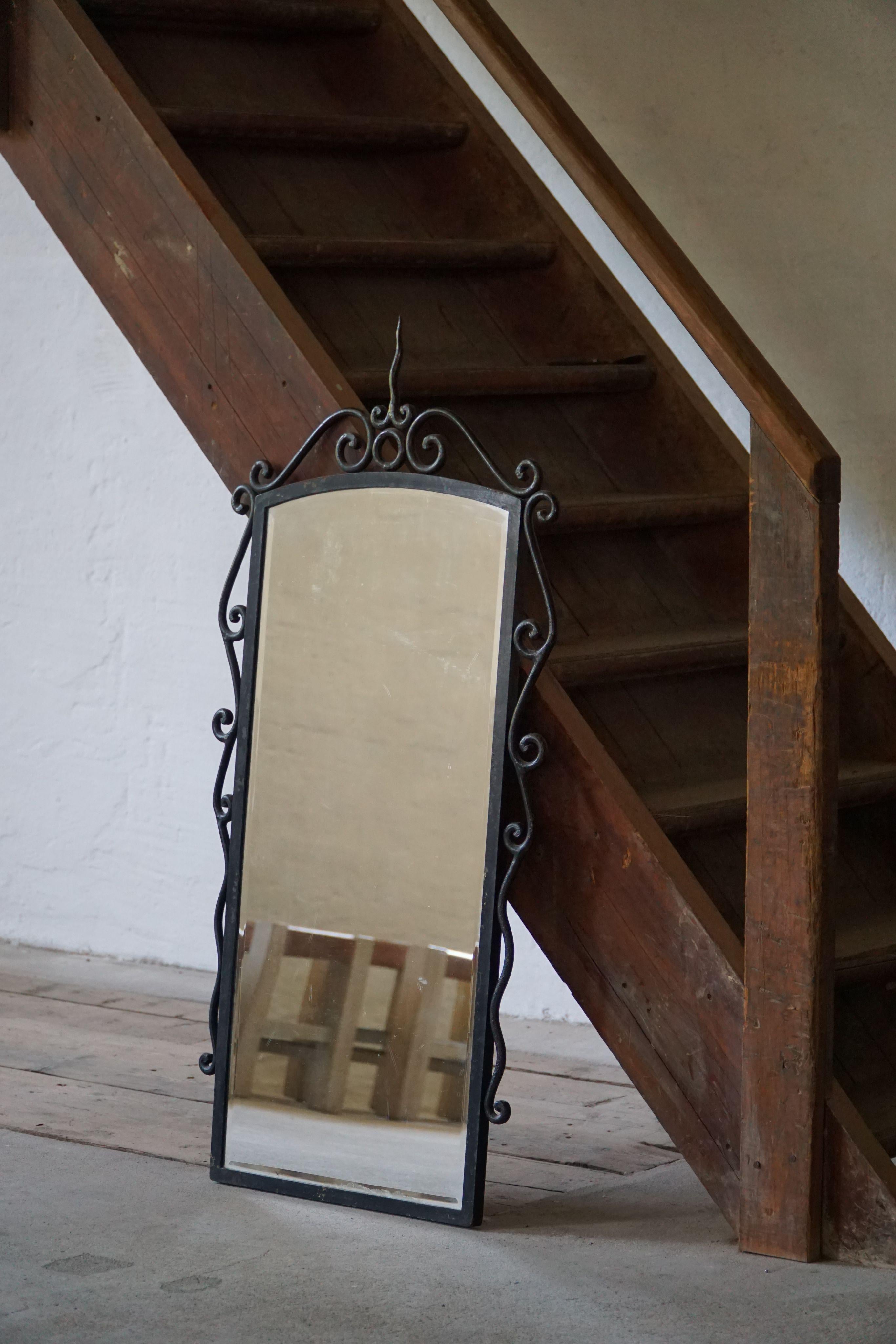 This decorative mirror is framed in dark wrought iron, shaped with gentle curves and crowned by an elegant pointed finial — a refined, almost architectural detail that gives the piece a romantic and graphic silhouette. Made in France in the