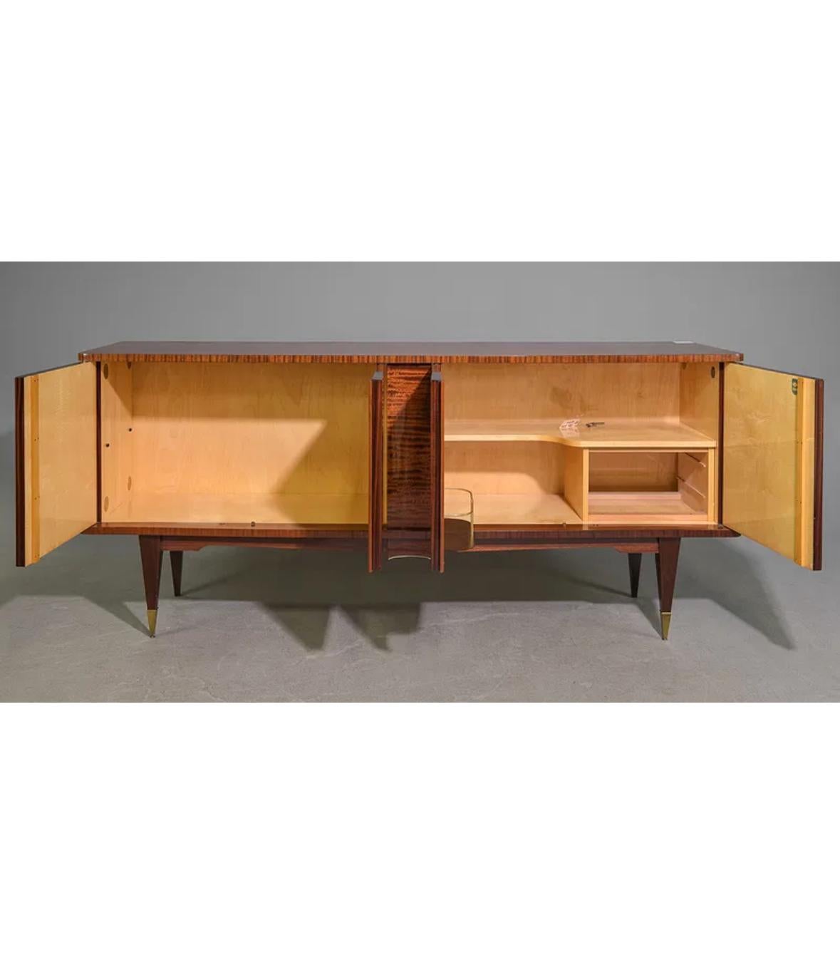 This sideboard, or credenza, includes a sleek built-in bar cabinet and highlights the dramatic, patterned grain of Macassar wood. Generous shelving provides ample storage, while tall, tapered legs with brass accents lend a refined touch. The piece