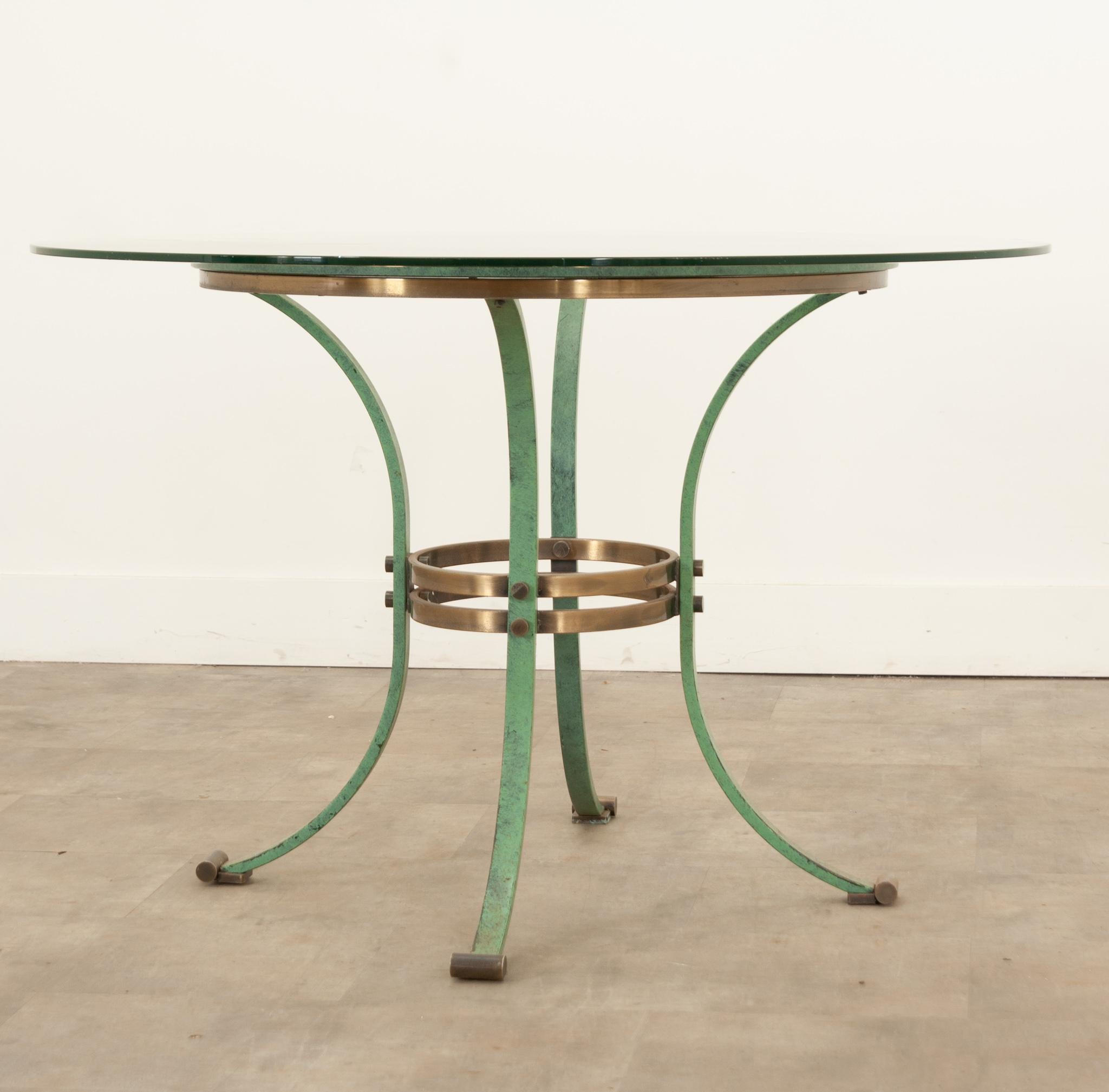 An eye-catching French Mid-Century pedestal dining table with removable beveled glass top, painted patina finish, and brass pedestal base. A beautiful round glass top with a beveled edge is supported by a circular brass pedestal base connected to