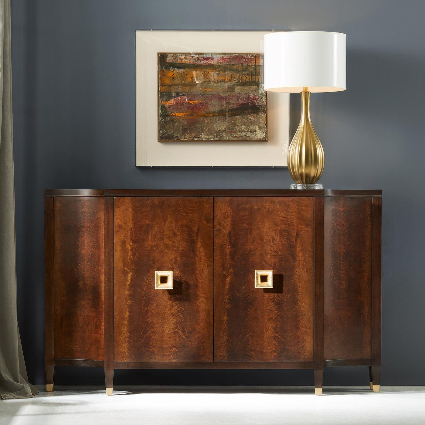 La credenza mette in mostra l'eleganza naturale del legno di noce lavorato abbinato a una raffinata ferramenta in foglia d'oro. L'impiallacciatura in noce abbinata crea un motivo scenografico su tutte e quattro le ante, mentre le maniglie squadrate