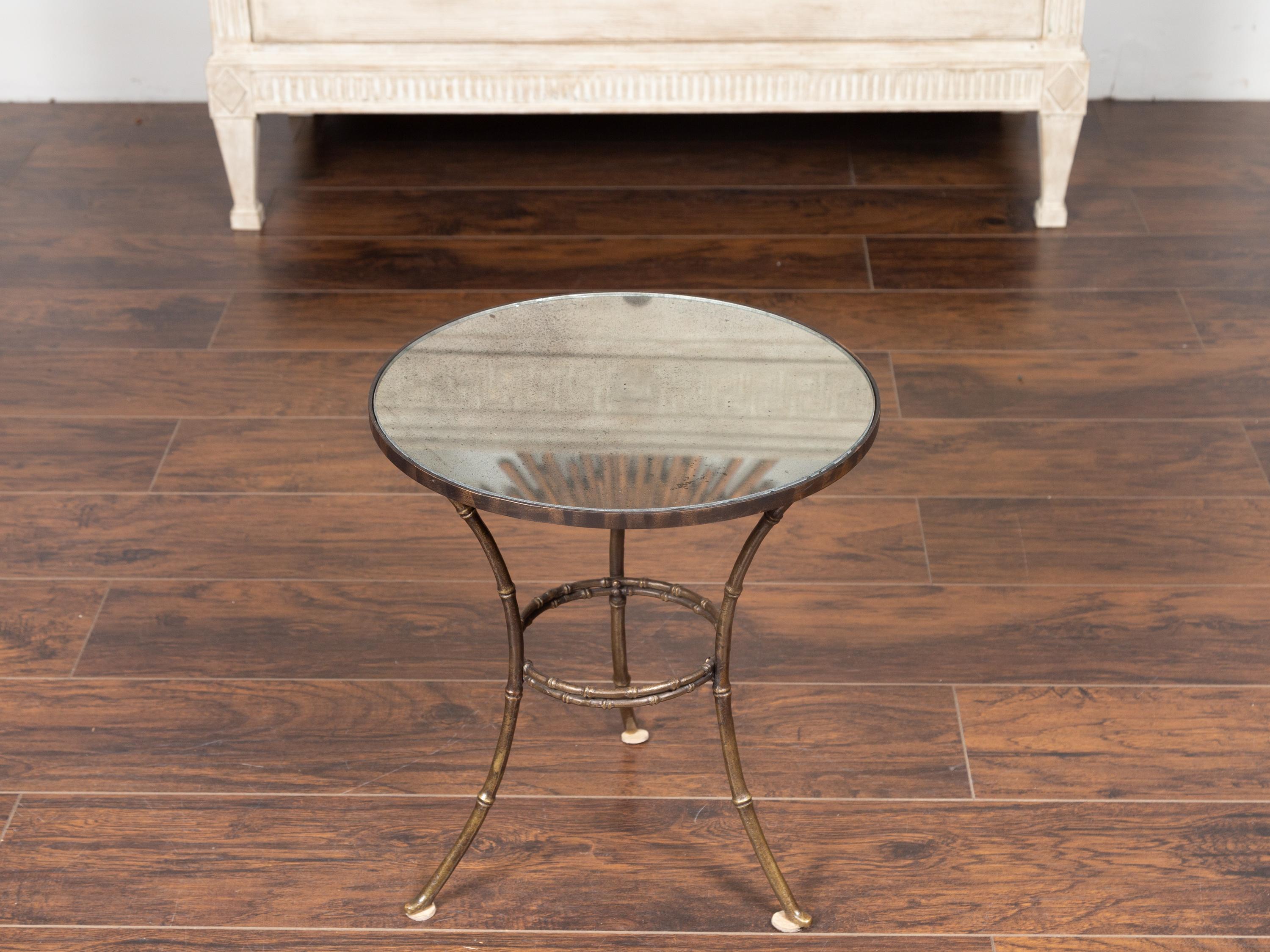 A French brass faux-bamboo style side table from the mid-20th century, with mirrored top. Born in France during the midcentury period, this lovely side table features a circular mirrored top with medium antiquing, sitting above a stylish brass