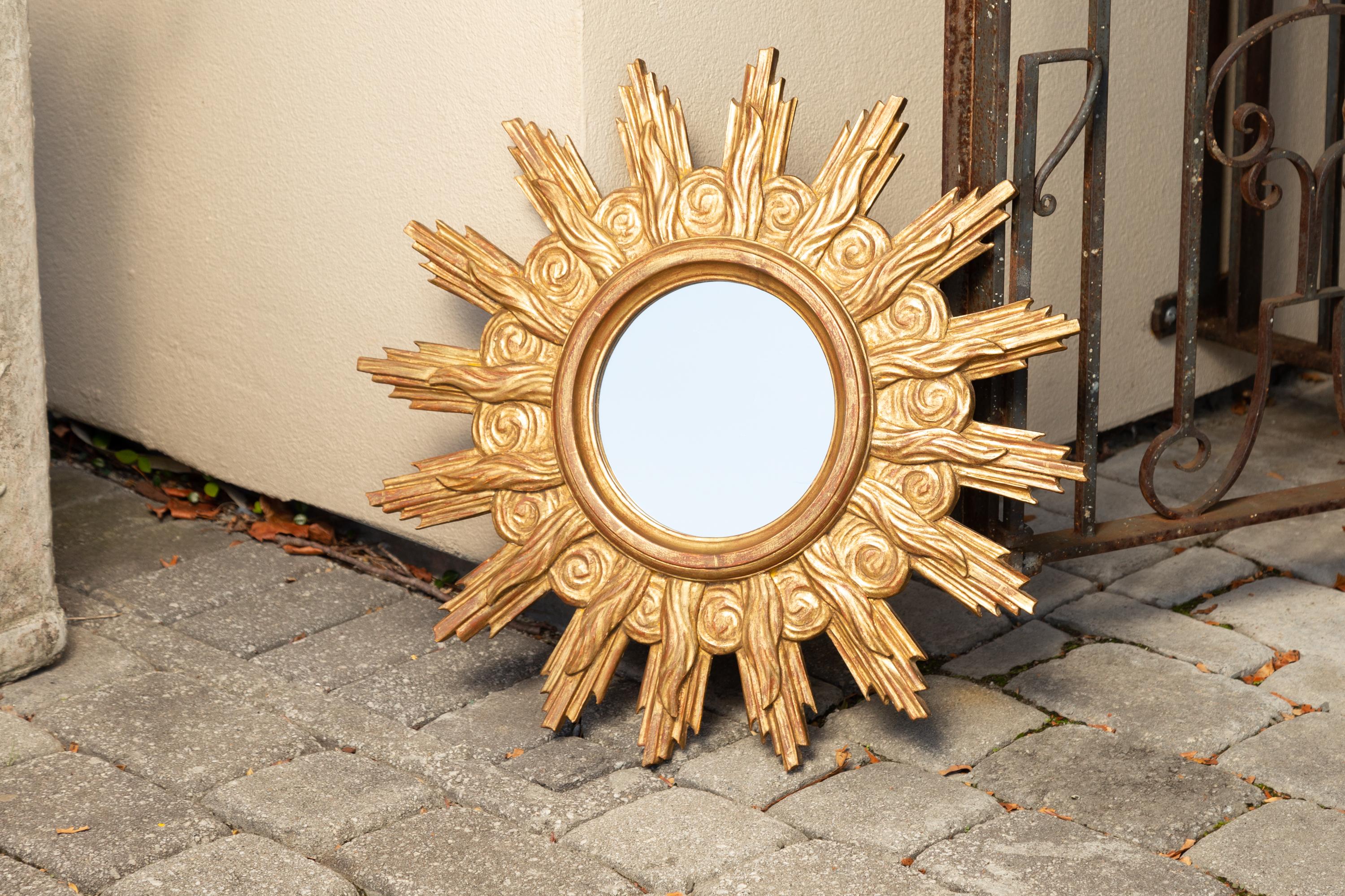 A vintage French carved giltwood sunburst mirror from the mid-20th century, with cloudy motifs and rays of varying sizes. Born during the midcentury period, this French sunburst mirror features a circular flat mirror plate, surrounded by an