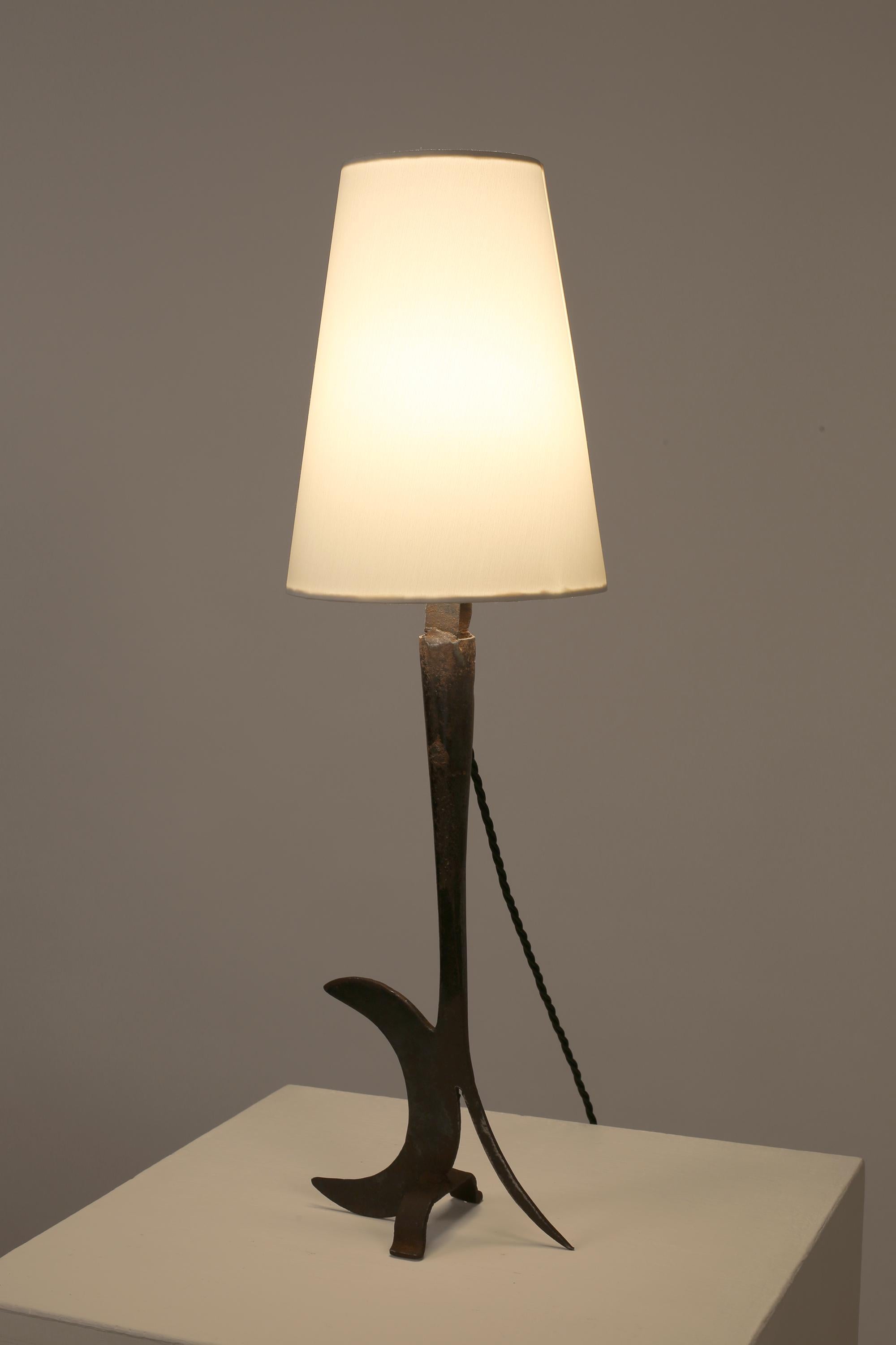 An abstract forged and welded iron table lamp, partly formed from an antique agricultural tool - typical folk-art work of the Provence region. French, c. 1950s. Supplied with a bespoke white cotton cone shade.