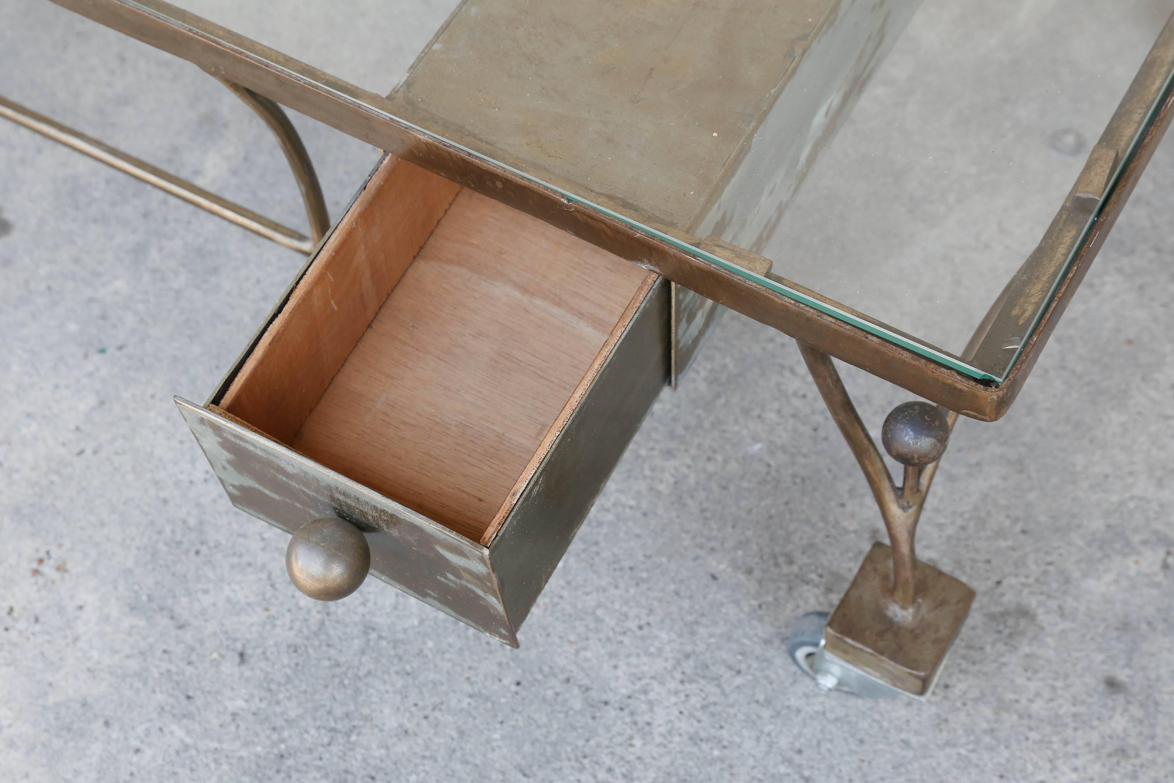 This is a darling midcentury French metal and glass cocktail table on wheels. The design of this piece even includes a narrow drawer at one side with a pull that echoes the decorative spherical accents on each leg. The finish of the metal is a