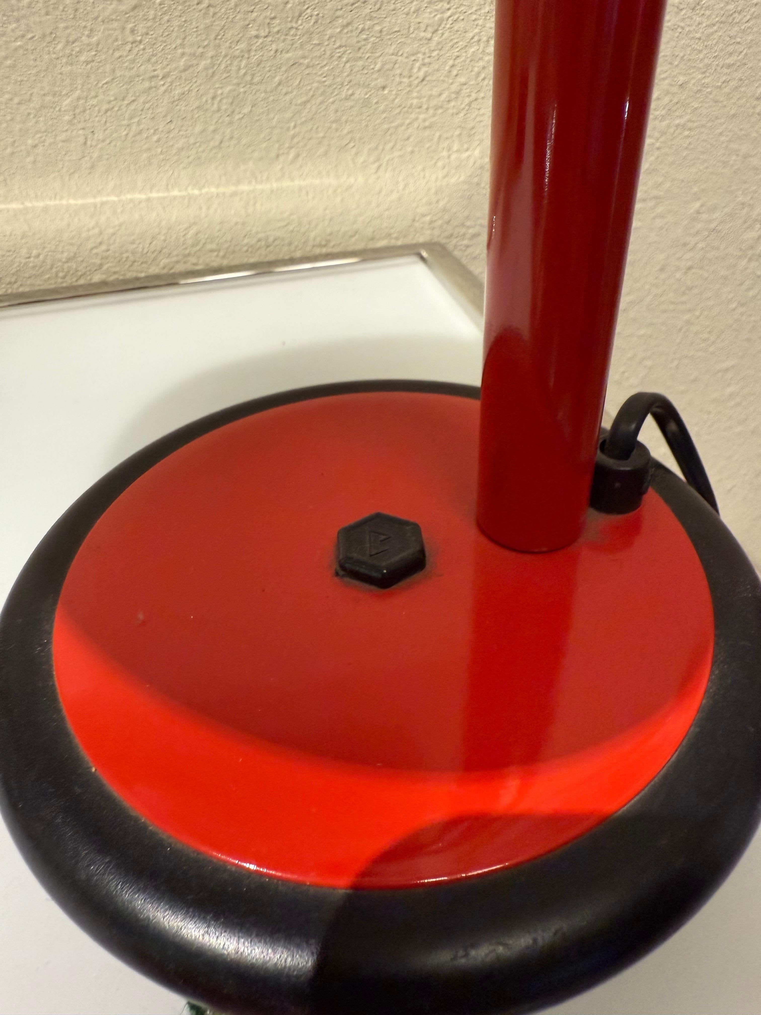 Diese Schreibtischleuchte von Aluminor verbindet funktionale Effizienz mit einem kühnen, modernistischen Stil. Ihr halbkugelförmiger roter Metallschirm kontrastiert stark mit dem schwarzen, perforierten Hals, wodurch ein dynamisches Spiel von Farbe