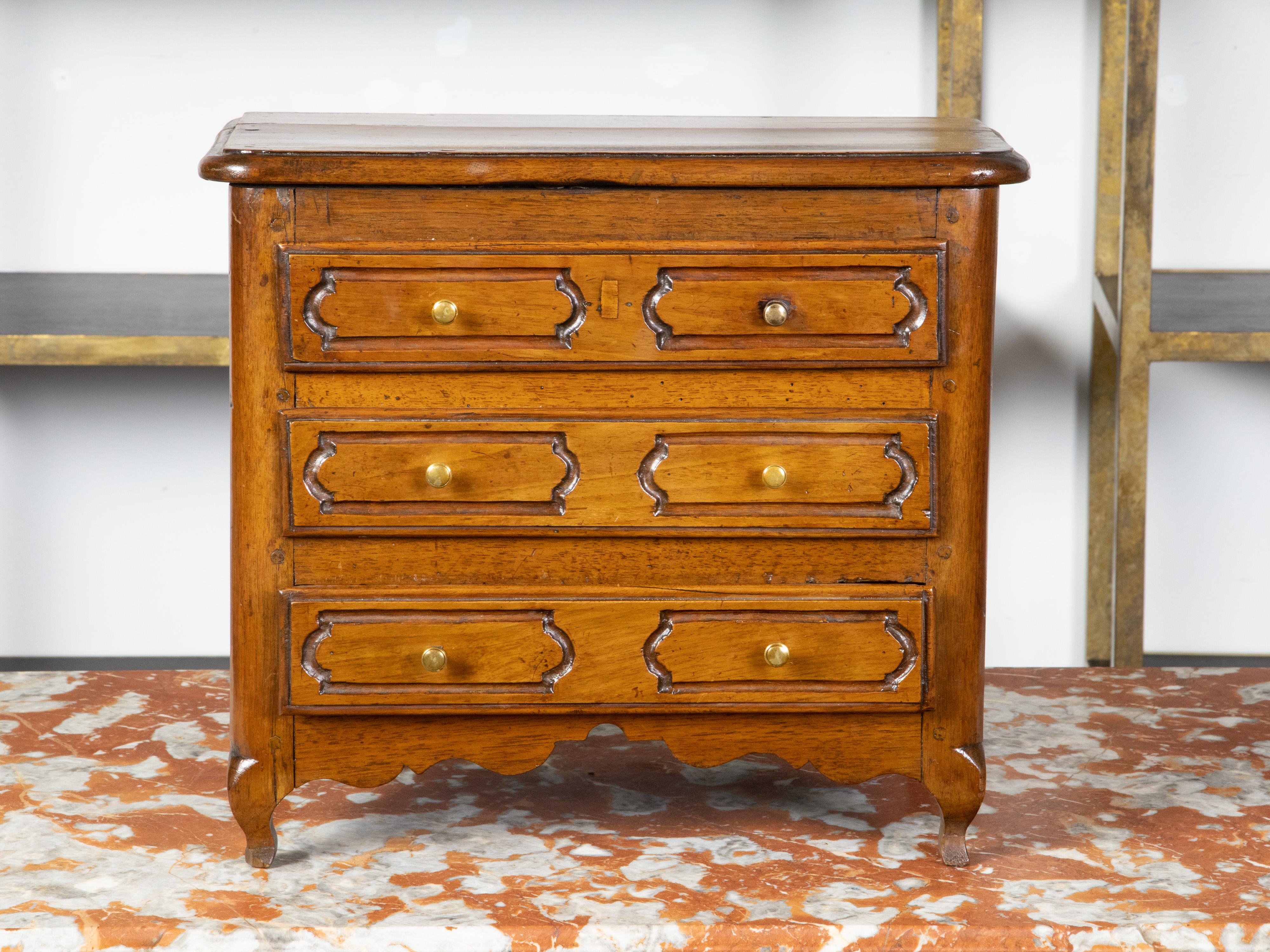 Una miniatura francese del XIX secolo in noce, costruita con lo stesso vocabolario dei mobili di provincia in scala reale. La cassa è sagomata con un piano dolcemente arrotondato, angoli smussati e un grembiule smerlato che poggia su corte gambe